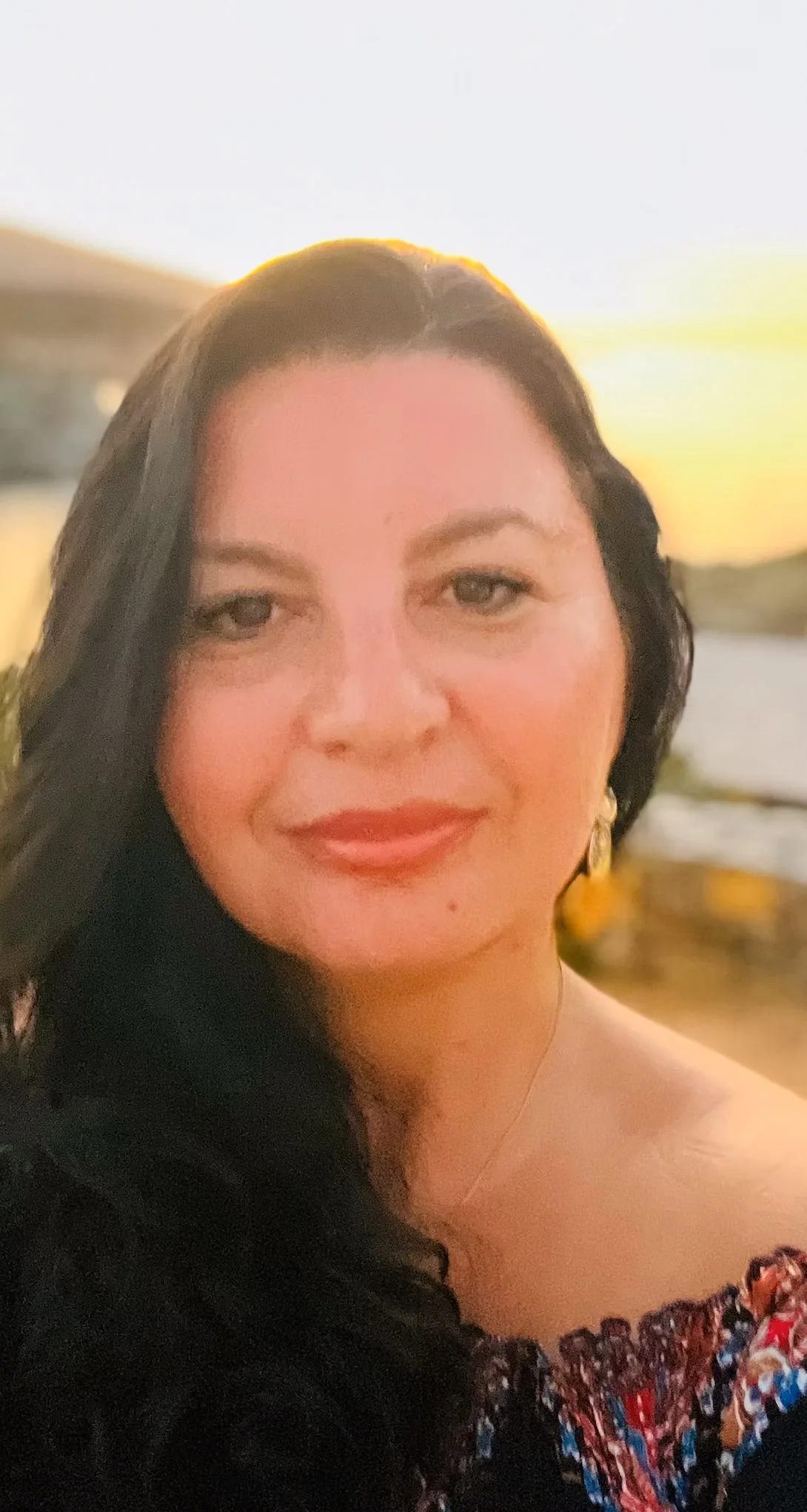 A woman with black hair and fair skin, wearing gold earrings and a colorful top, smiling with a sunset in the background.