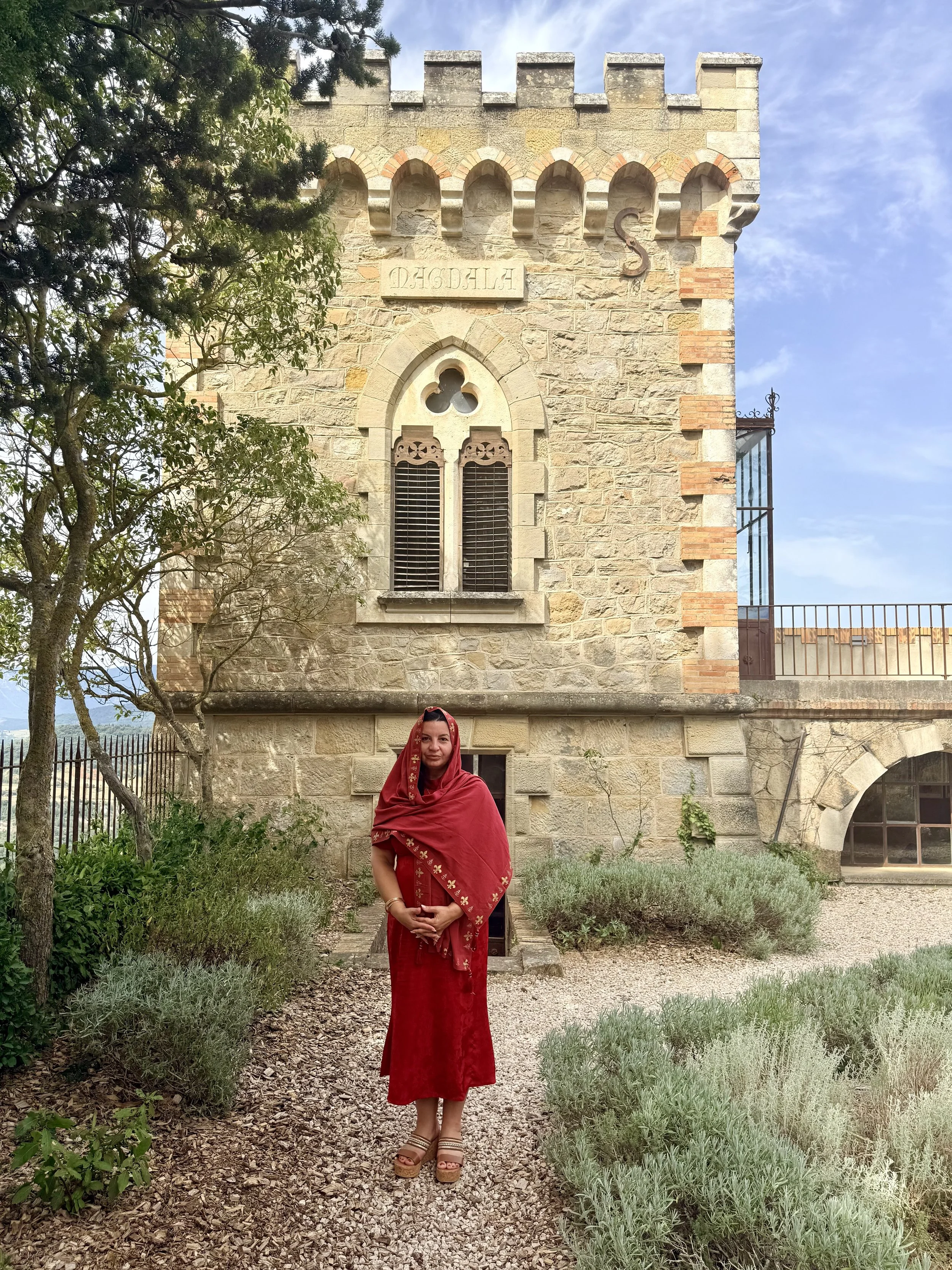 A woman dressed in a red sari and traditional jewelry stands in a garden in front of a stone building with arched windows and decorative brickwork.