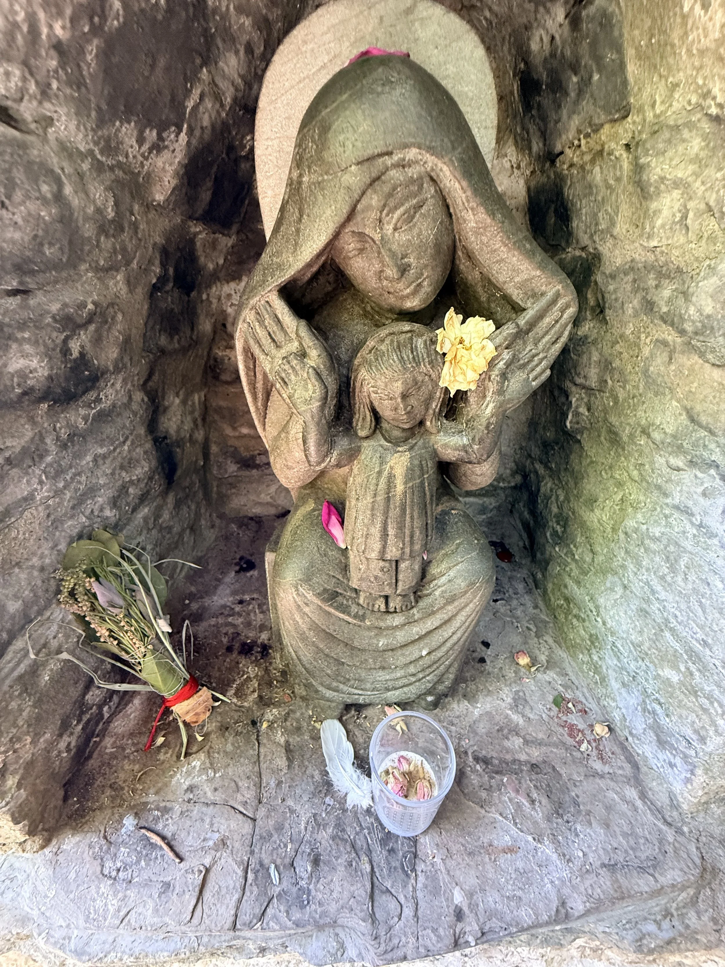 A stone sculpture of a woman holding a baby, with flowers and offerings placed around it, located inside a small cave or niche.