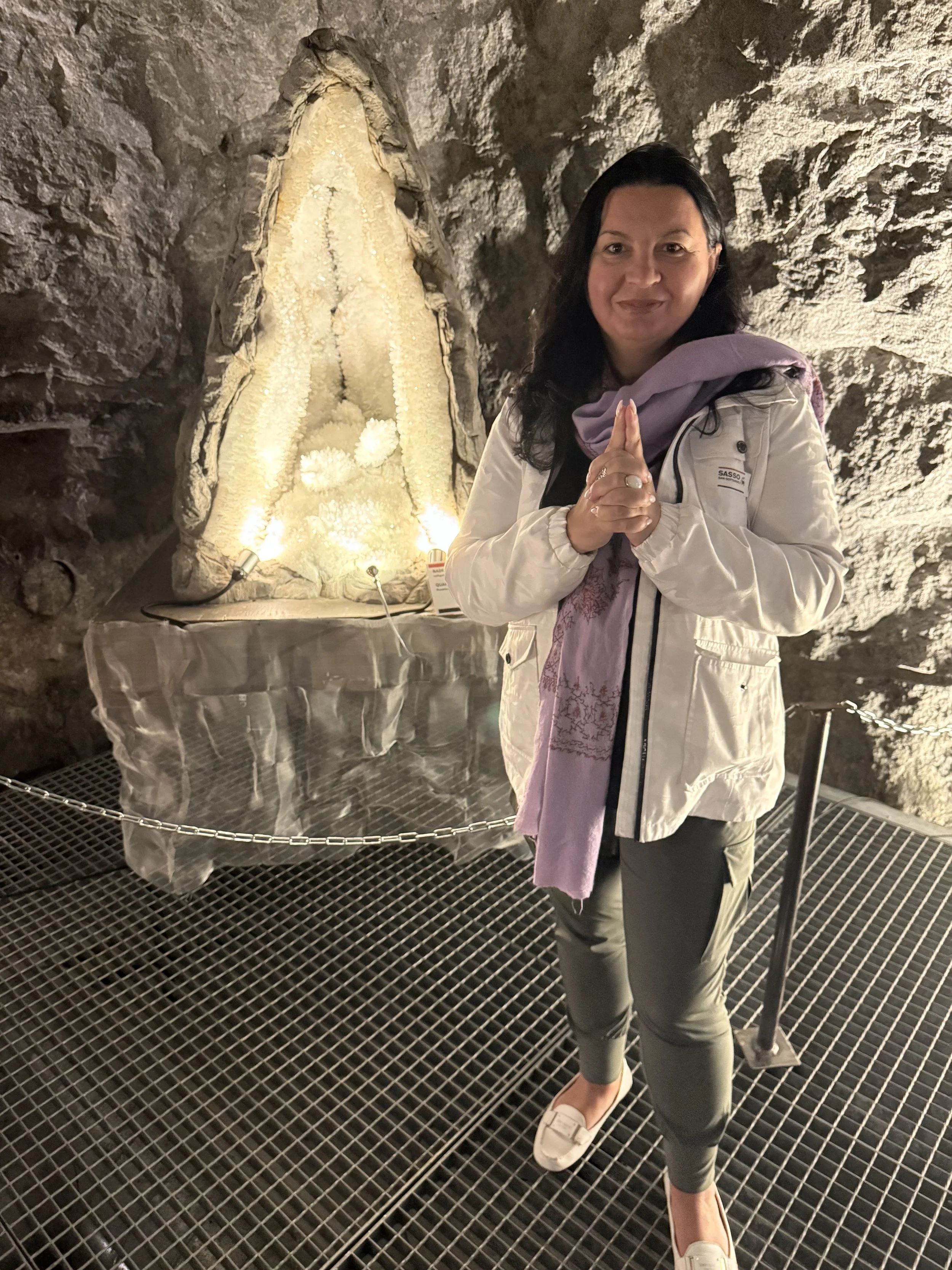 A woman with dark hair, wearing a white jacket, purple scarf, and gray pants, standing on a metal grid floor inside a cave or underground site, with her hands pressed together in a prayer gesture in front of a large stalactite formation.