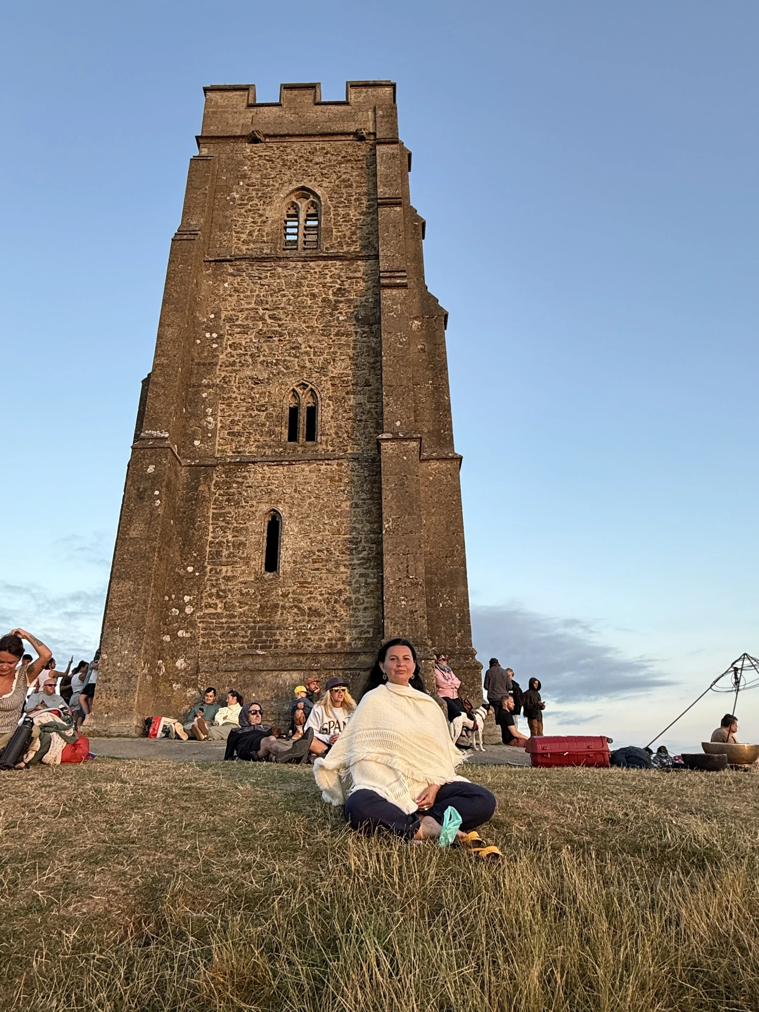 People sitting and relaxing on grass in front of a tall brick tower during sunset.