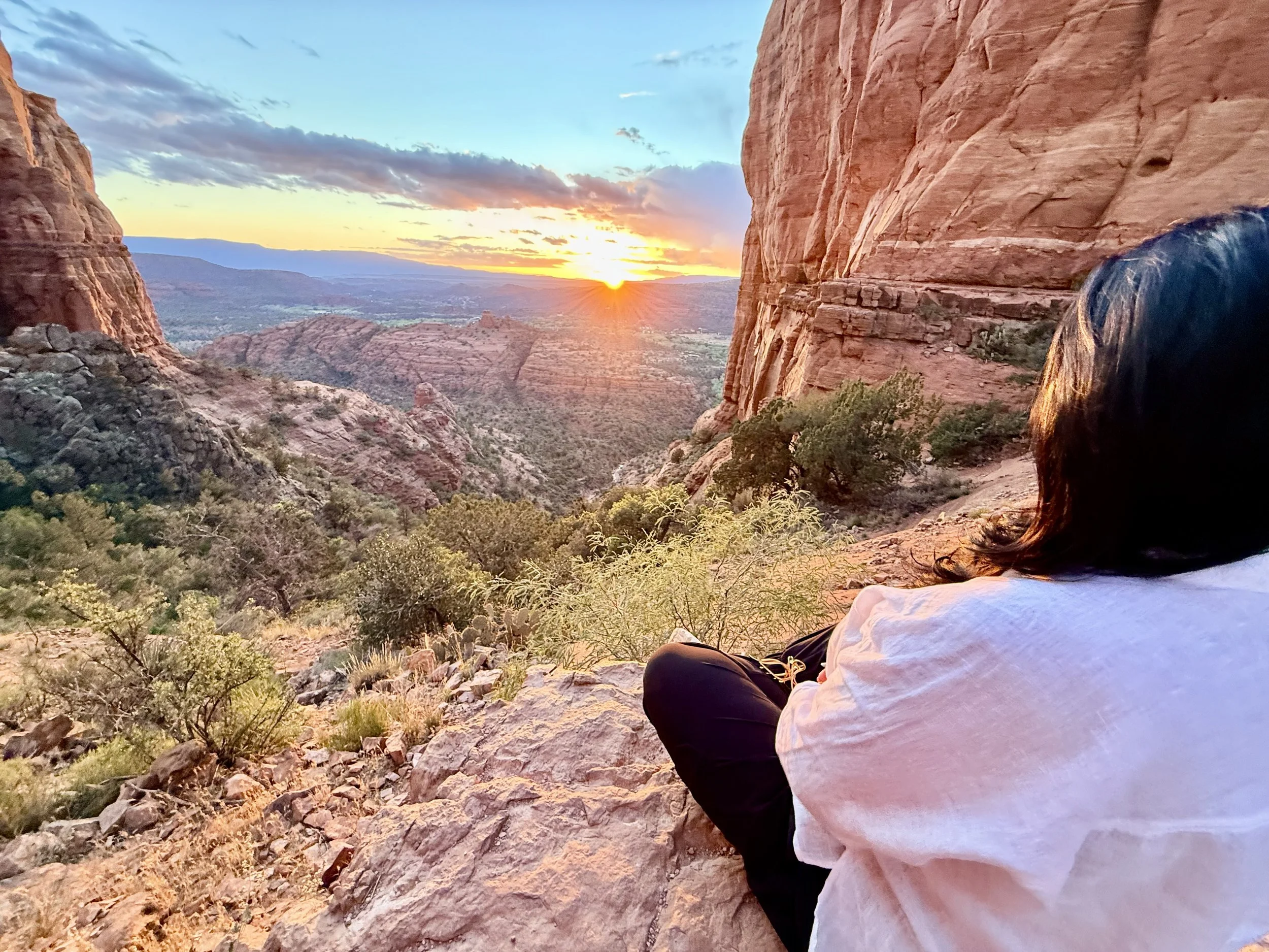 A person wearing a white shirt and black pants is sitting on rocky terrain, facing a scenic canyon with red rock formations at sunset, with the sky partly cloudy.