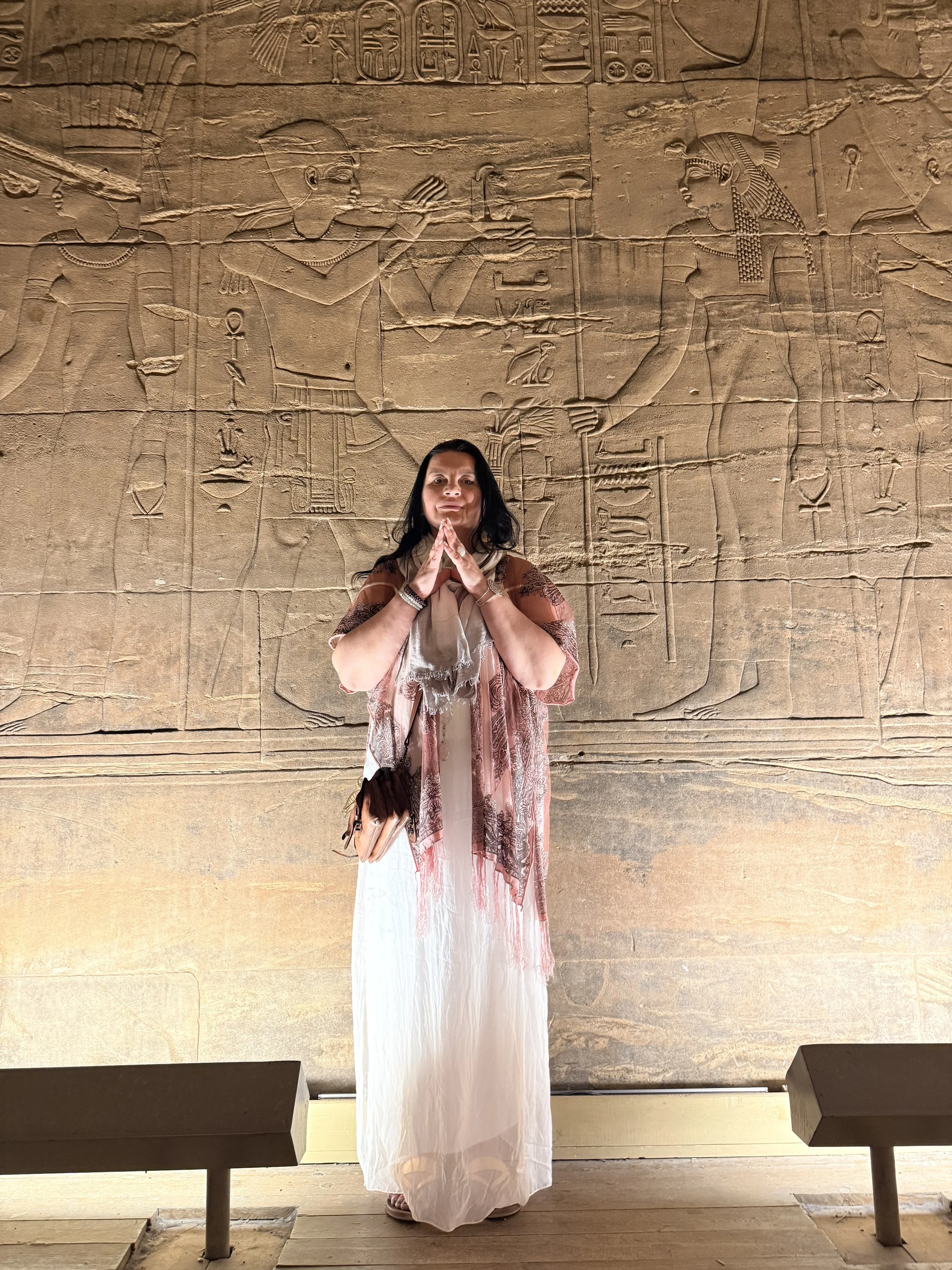 A woman standing in front of ancient Egyptian wall carvings, wearing a long light-colored dress and a patterned shawl, with her hands in a prayer position.