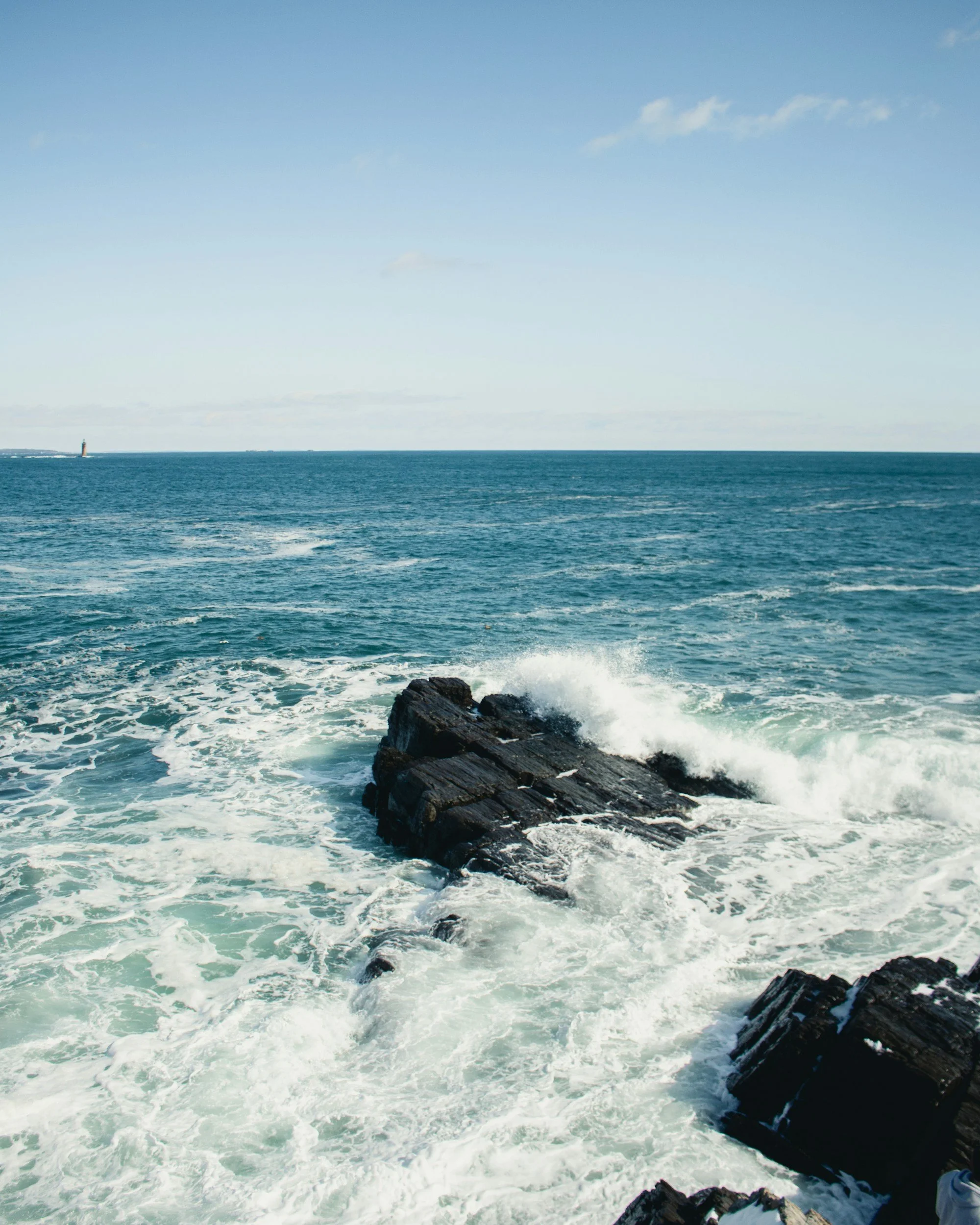 Ocean waves crashing against dark rocks under a clear blue sky with a lighthouse visible in the distance.