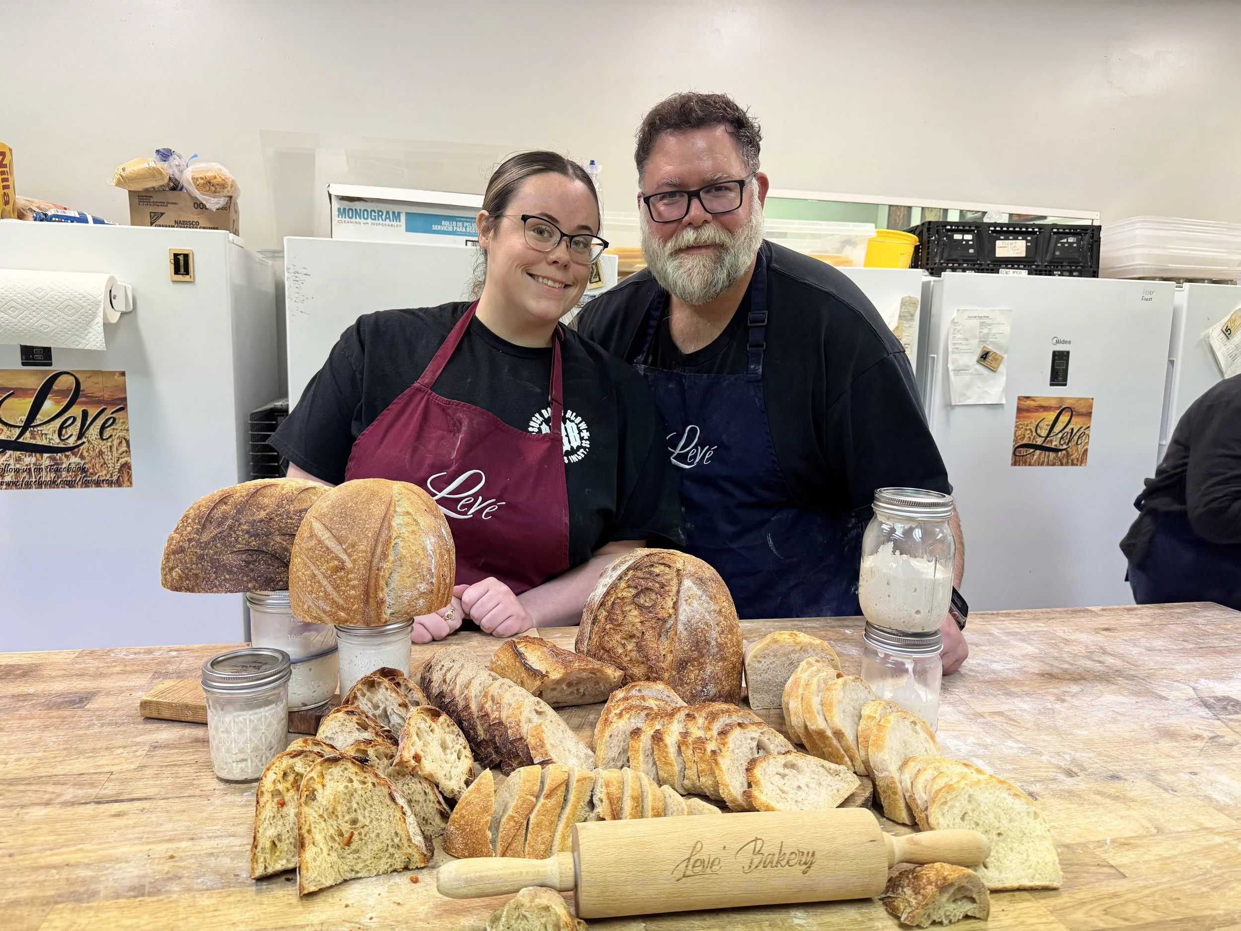 A man and woman wearing black shirts and aprons with 'Léve' written on them, standing behind a wooden table with various loaves of bread and baked goods, in a kitchen or bakery setting.