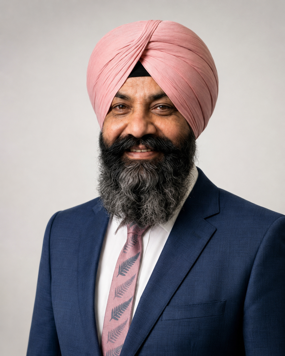A man with a pink turban, black beard with gray streaks, dressed in a dark blue suit, white shirt, and a pink tie with gray fern patterns, smiling at the camera against a plain background.