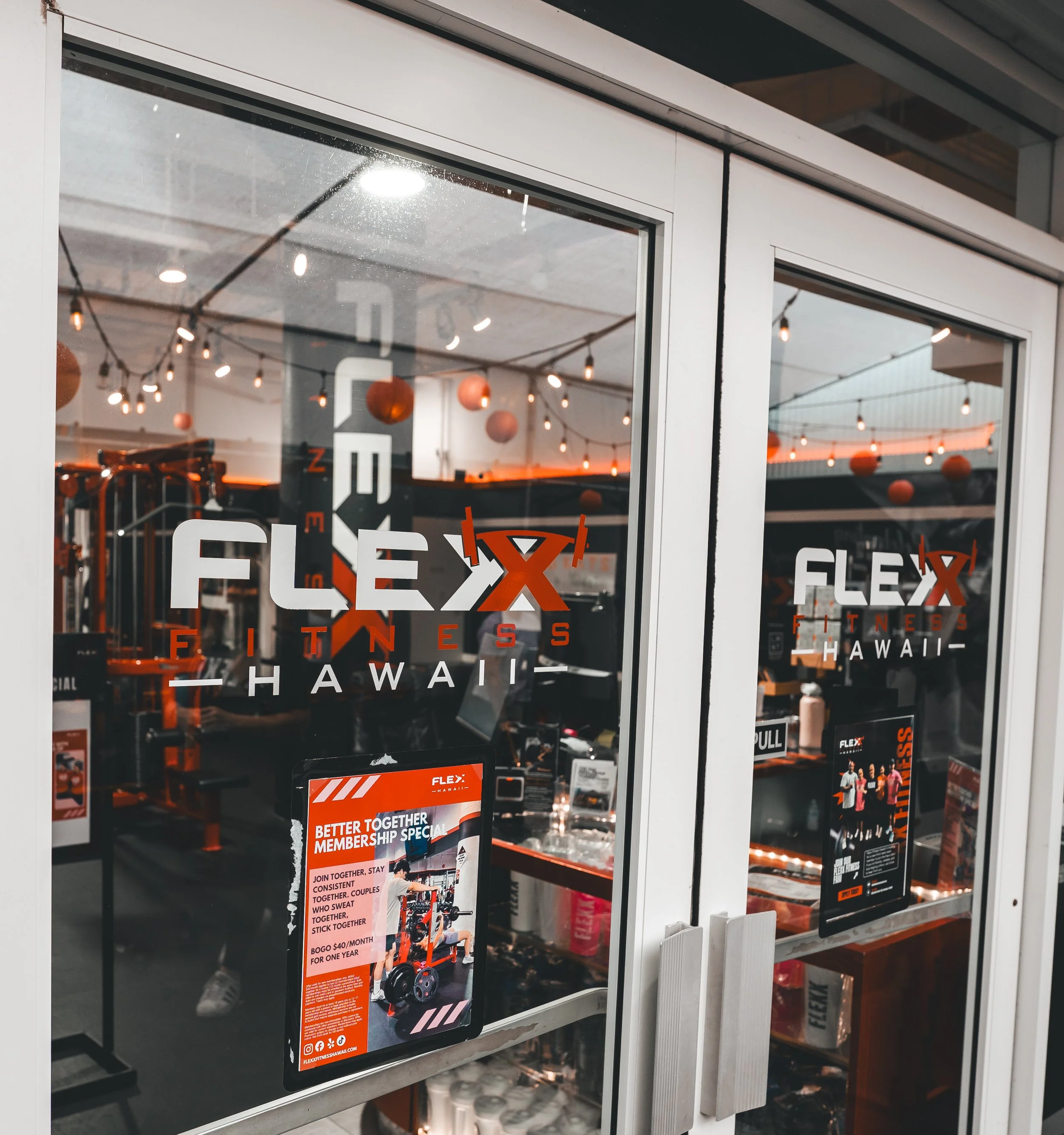 Store window with signs and decorations inside, including hanging lights and red lanterns, advertising a fitness center called Flex Fitness Hawaii.
