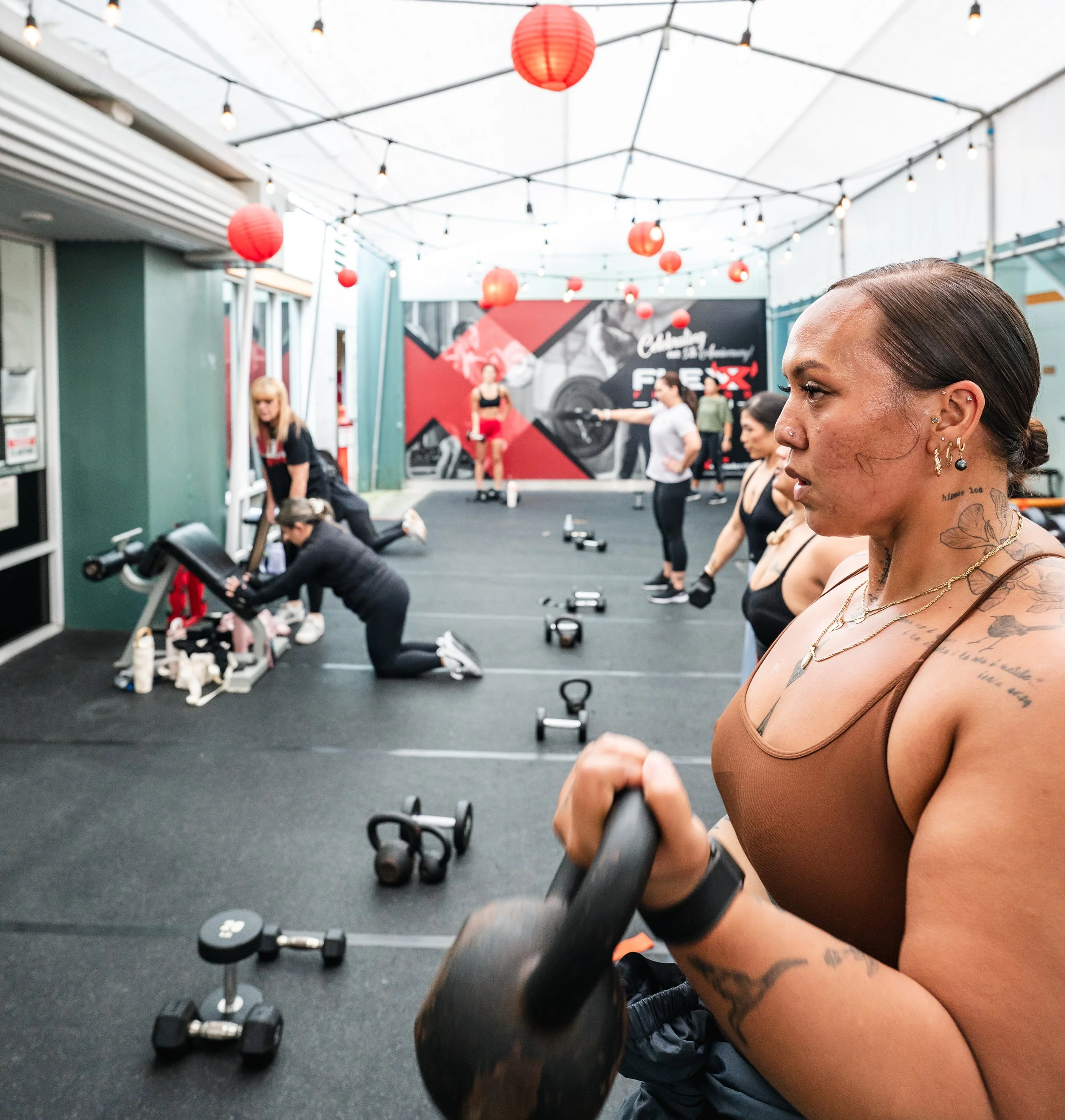Women exercising in a fitness class in an outdoor gym area with hanging lights and red paper lanterns, some women are lifting weights or doing other workouts.