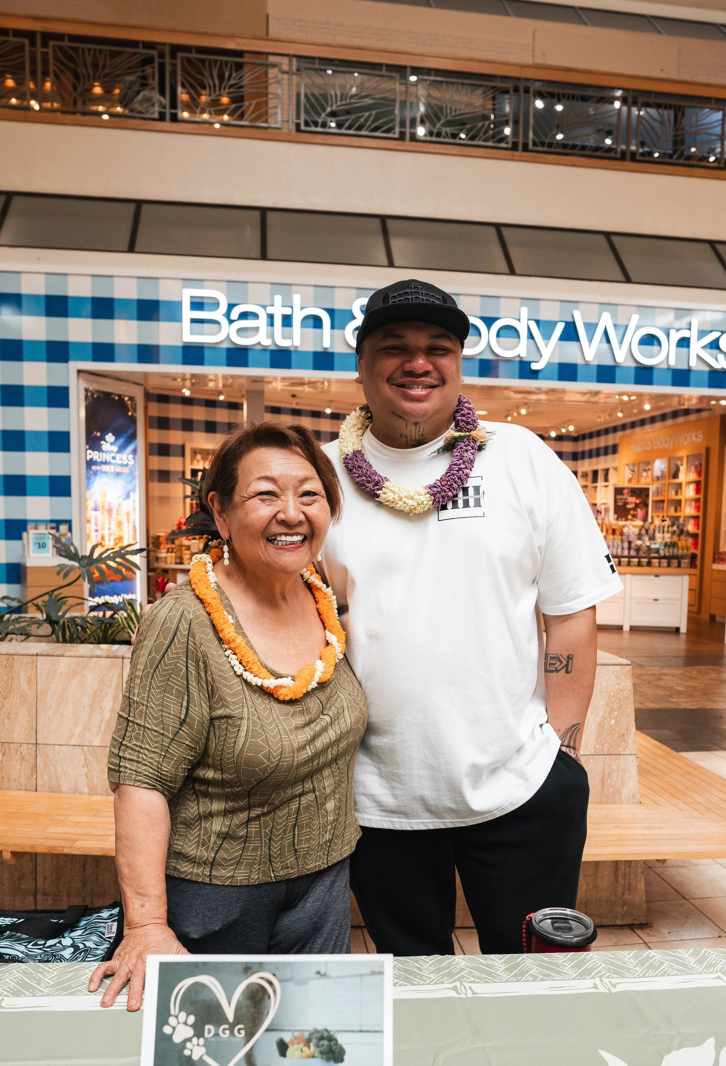 A woman and a man standing together inside a shopping mall in front of a Bath and Body Works store, smiling at the camera, both wearing leis made of flowers.