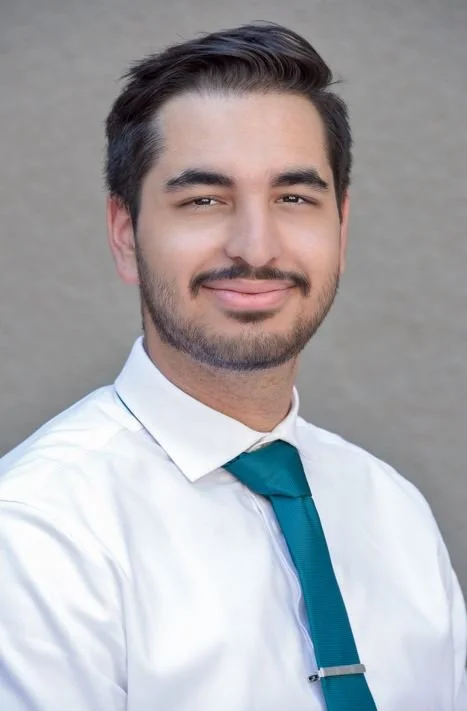 Peter La Mazza a young man with dark hair and a trimmed beard, wearing a white shirt and teal tie, smiling and standing against a neutral background.