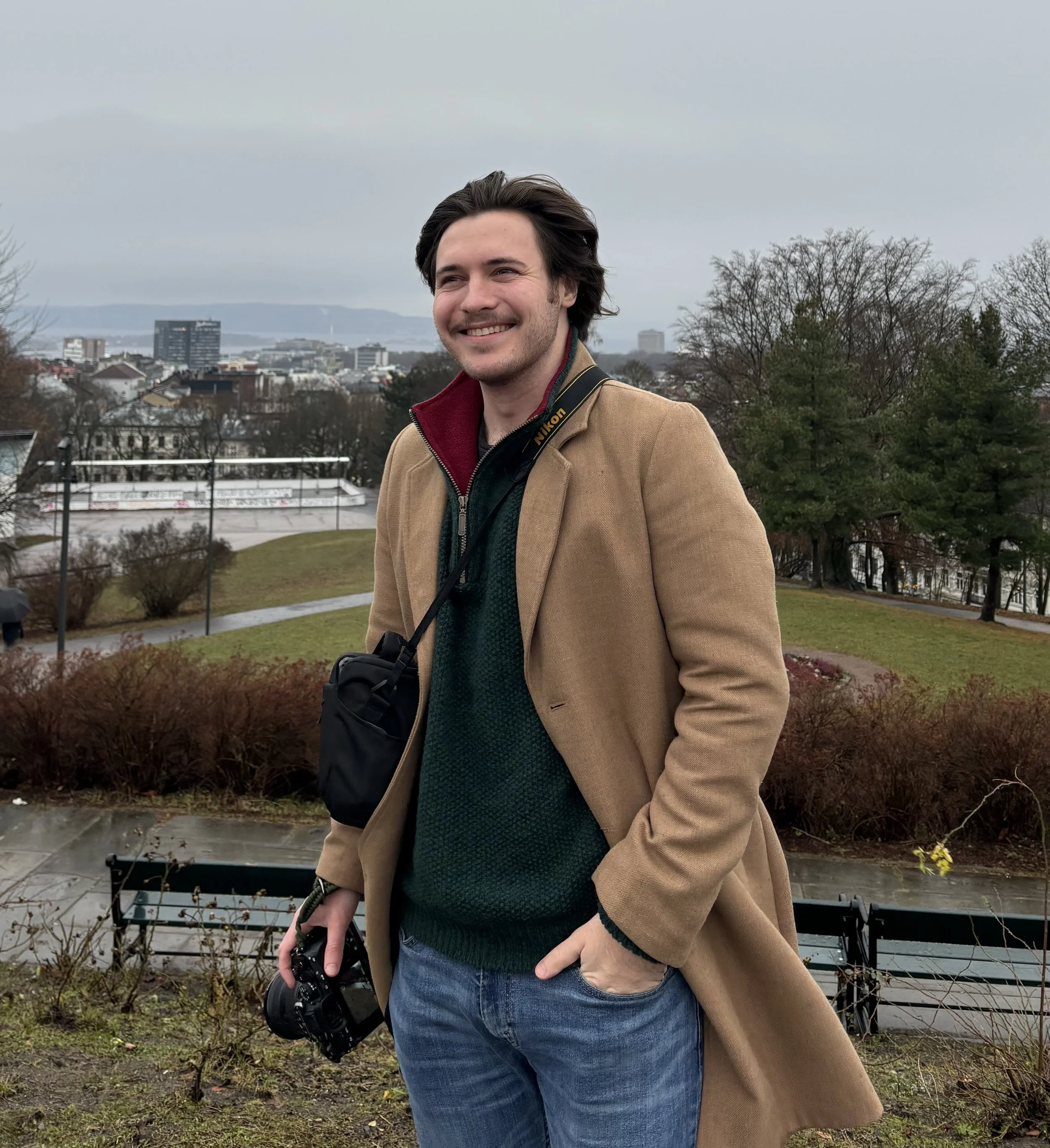 A young man with dark hair smiling outdoors, holding a camera, wearing a tan coat over a green sweater, standing in a park with trees and a city skyline in the background on a cloudy day.