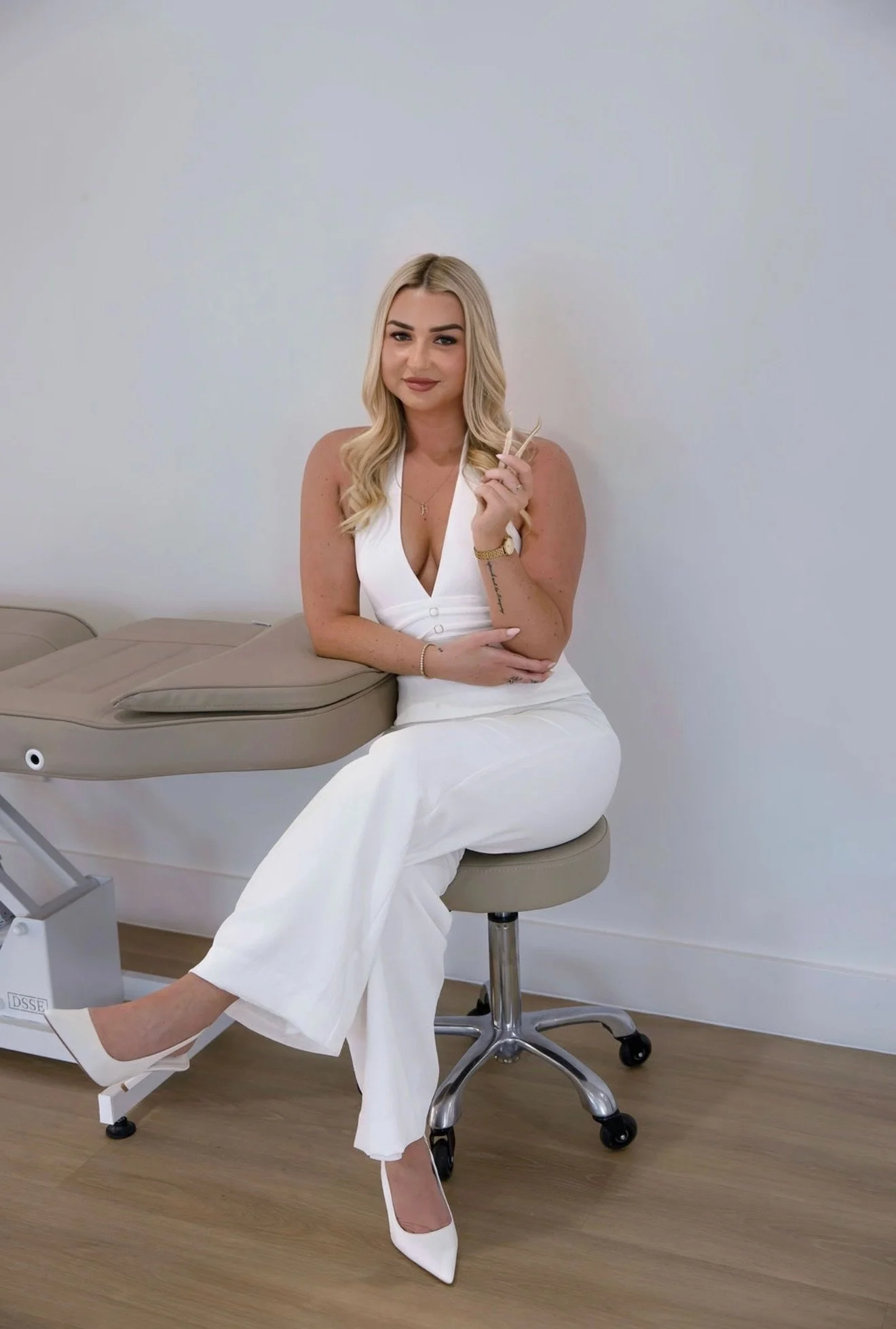 A young woman in a white sleeveless jumpsuit and white heels sitting on a beige examination table in a medical office, holding an object in her hand and smiling at the camera.