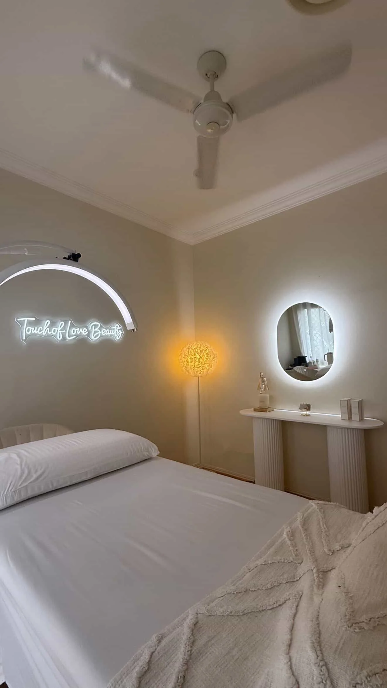 A bedroom with a white bed, a decorative pillow, a round mirror with backlit lighting, a small table with decor items, a neon sign that says 'Touch of Love Beauty,' a yellow floor lamp, and a ceiling fan.