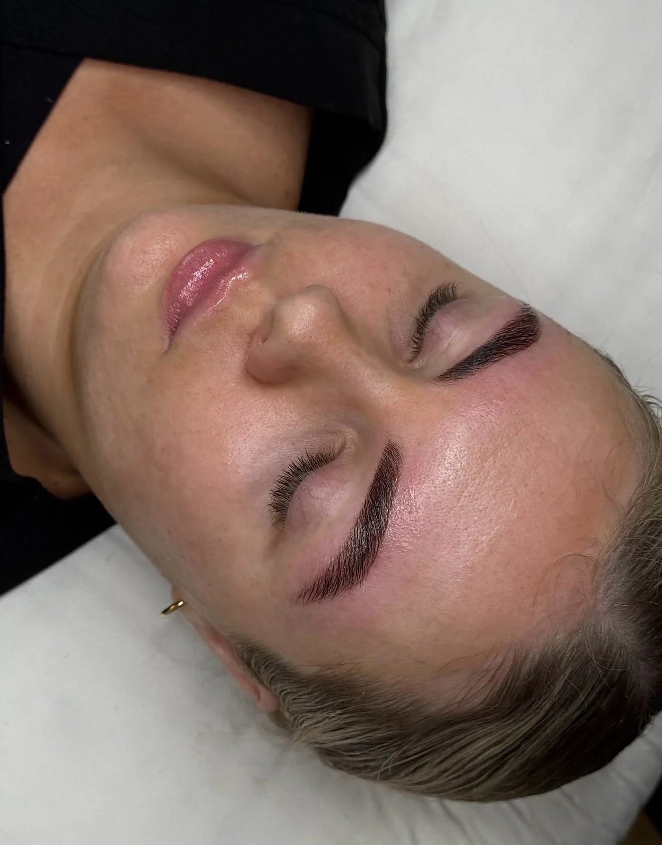 A woman lying down with freshly groomed eyebrows and clear skin, resting on a white surface, wearing a black top.