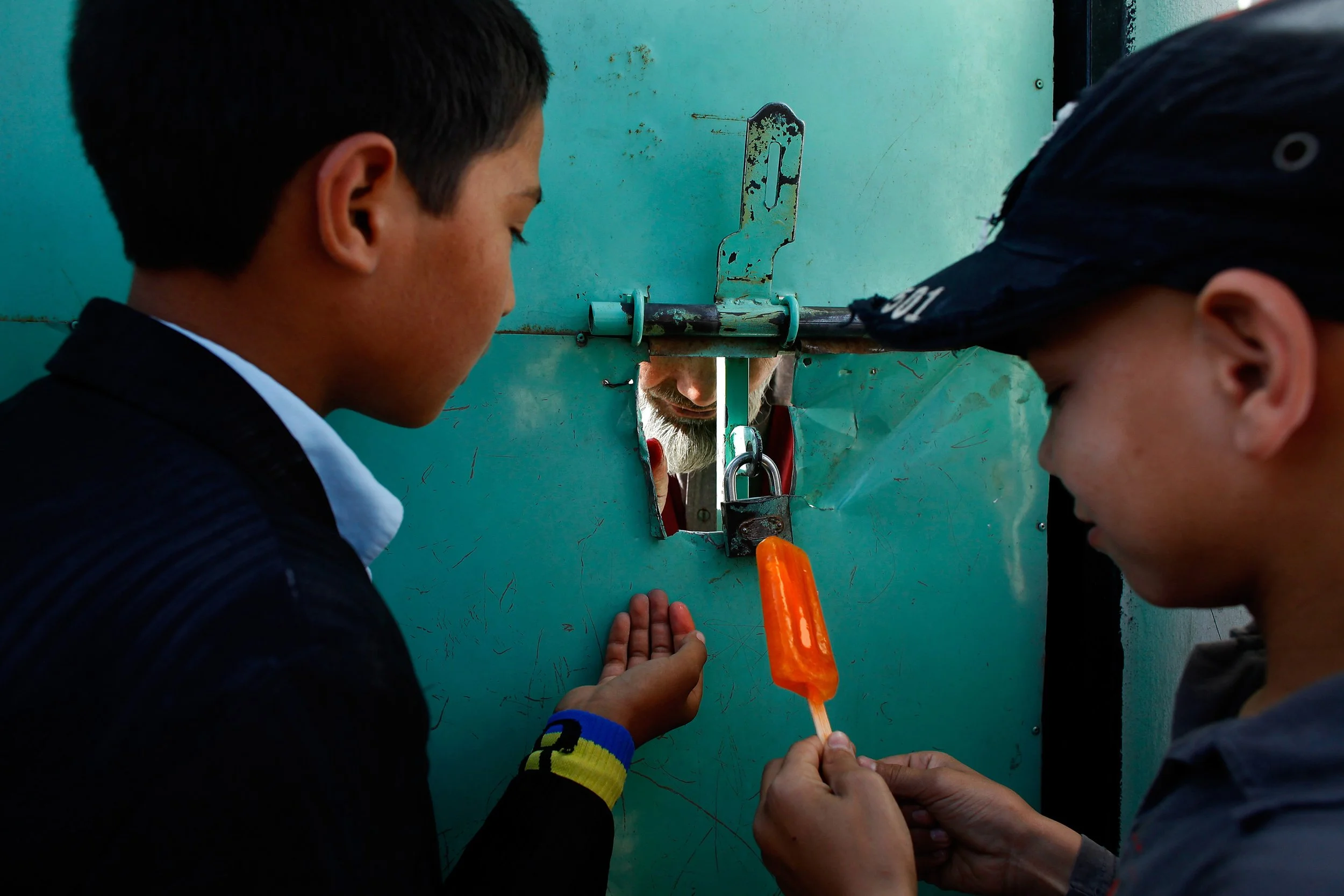 Students buy popsicles from a man selling them through the gate of a schoolyard. Boys and girls attend classes in separate shifts. Classrooms are crowded. Books supplies and teachers are scarce.