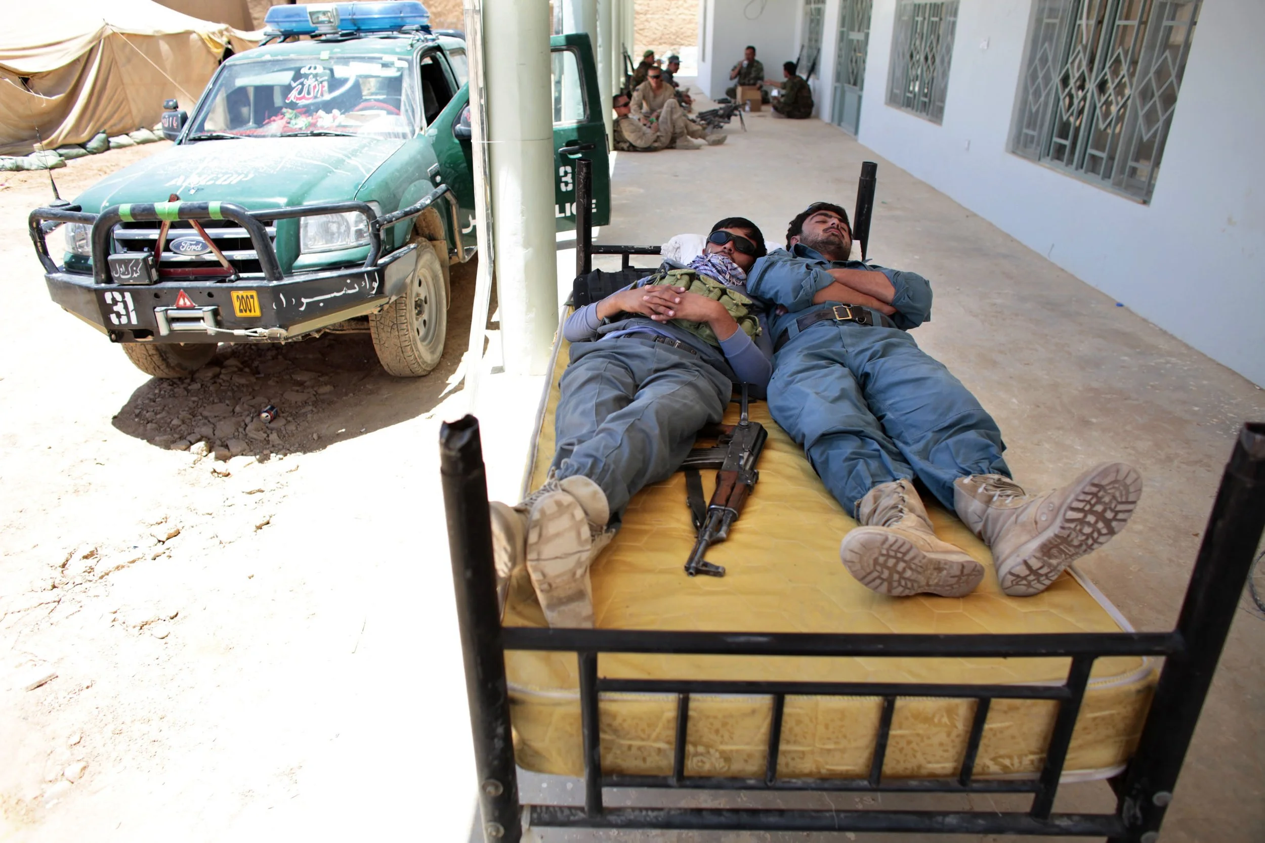 At the dedication of a new government center in Helmand, Afghan police officers nap on a bed inexplicably placed on the front porch. For coalition trainers, instilling discipline in local security forces remains a persistent challenge.