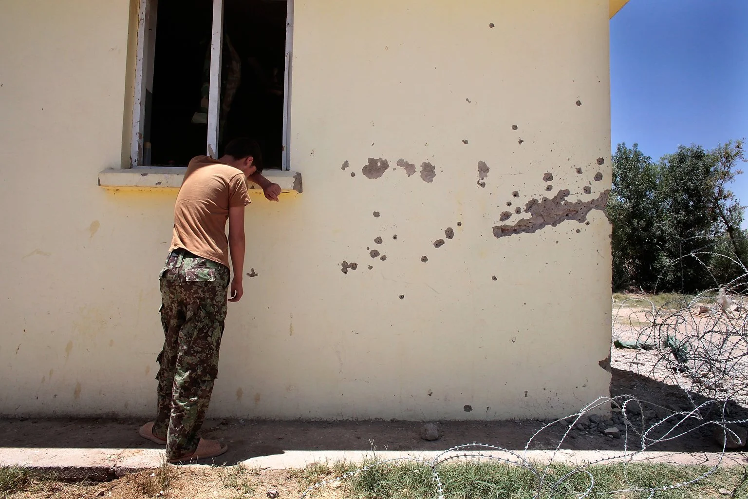 Exhausted and overwhelmed, a member of Afghanistan’s embattled security forces leans against the wall of a school in Marjah — his back resting beside bullet holes left by Taliban fighters. The scars of conflict surround him, visible on both the walls