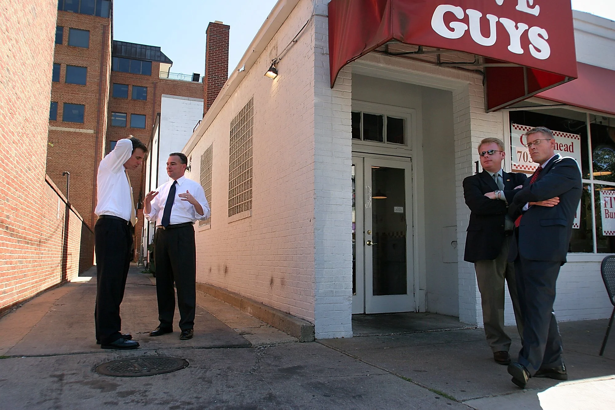 While an Executive Protection Unit officer and press secretary Kevin Hall secure the area, then–Virginia Gov. Mark Warner, far left, listens to Lt. Gov. Tim Kaine as they wait for their to-go burger order outside a Five Guys restaurant in Alexandria