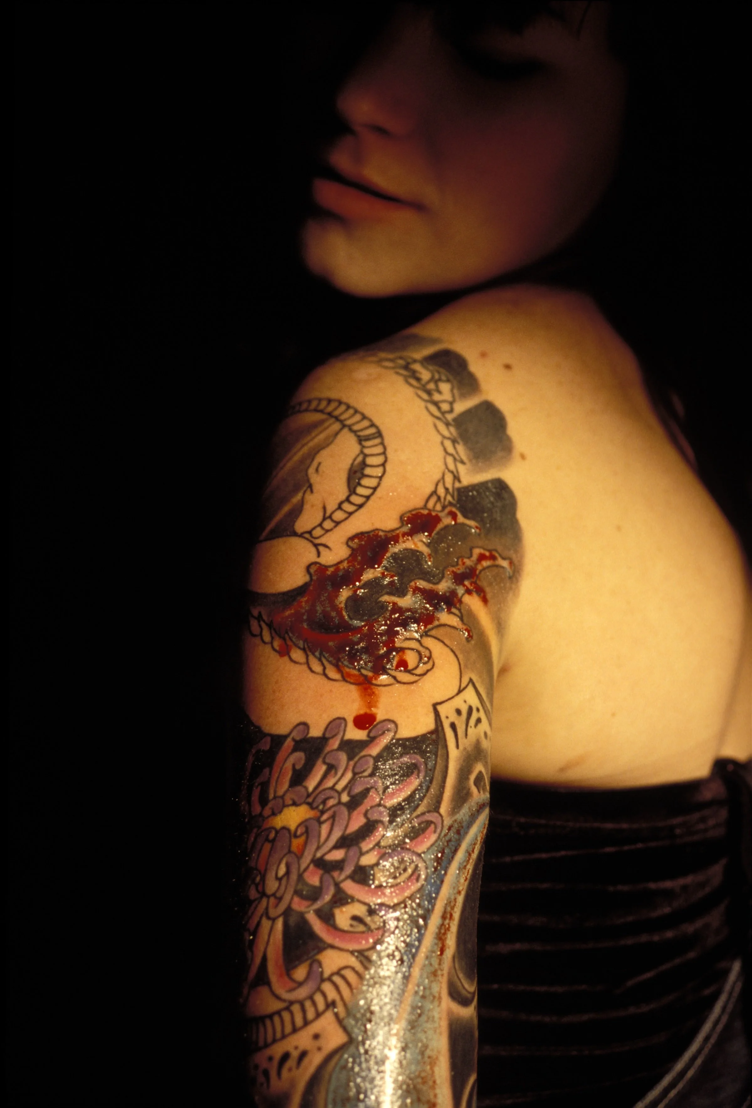 A woman poses for a portrait moments after receiving a fresh tattoo, its ink and blood still visible on the skin. The image captures the raw artistry and immediacy of body modification.