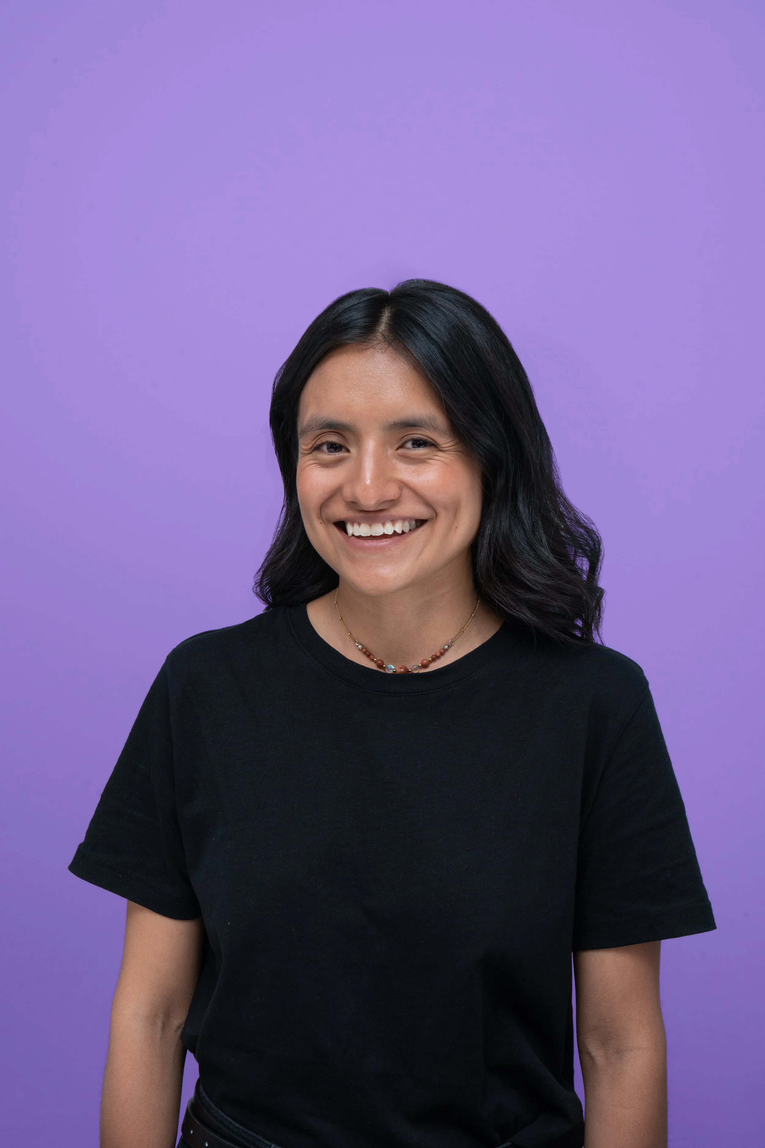Joven mujer sonriendo con fondo morado, cabello oscuro, camiseta negra y collar de cuentas.