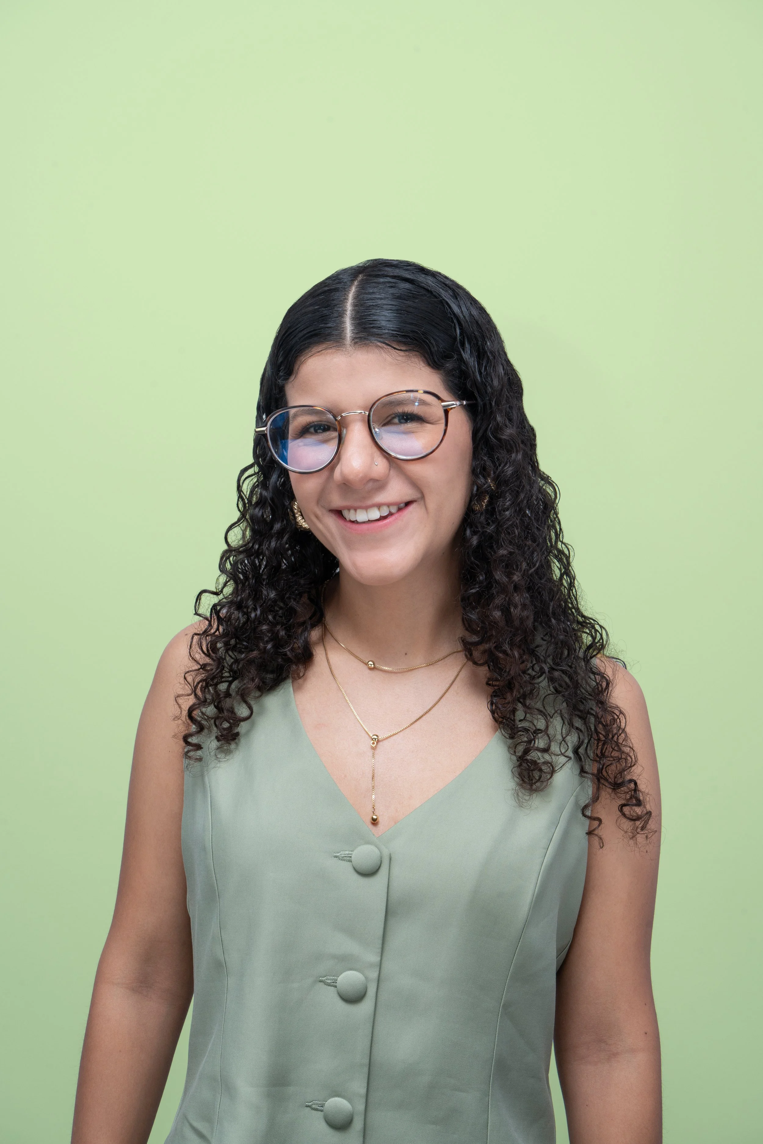 Juvenil mujer con cabello rizado, gafas y vestido verde claro, sonriendo, con fondo verde claro.