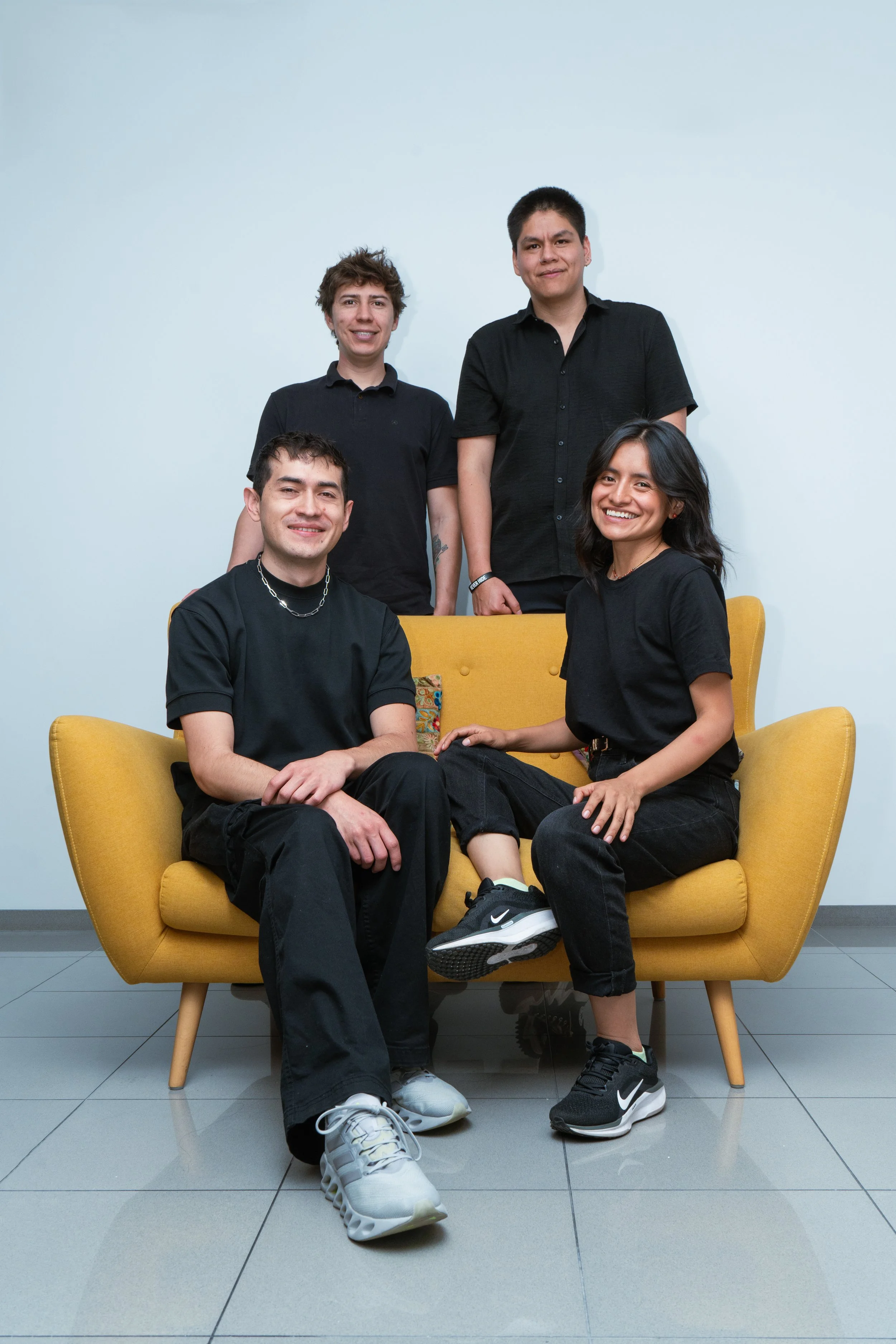 Cinco personas, tres hombres y una mujer, en una sala con fondo blanco, con dos personas en el sofá amarillo y tres de pie detrás, todos vestidos con ropa negra.