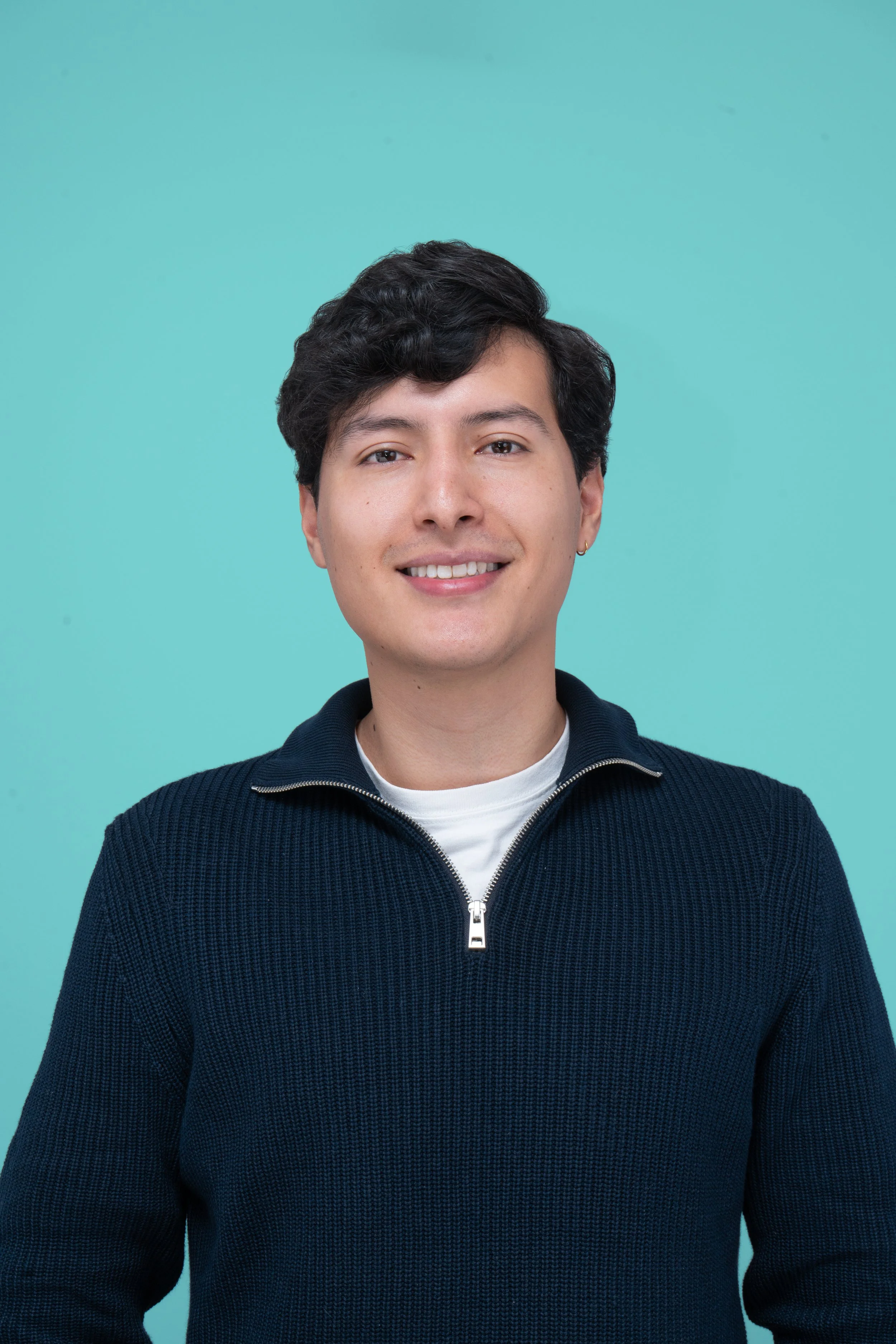 Retrato de un joven con cabello oscuro y ondulado, sonriendo, usando un suéter azul con cierre y una camiseta blanca debajo, frente a un fondo azul claro.