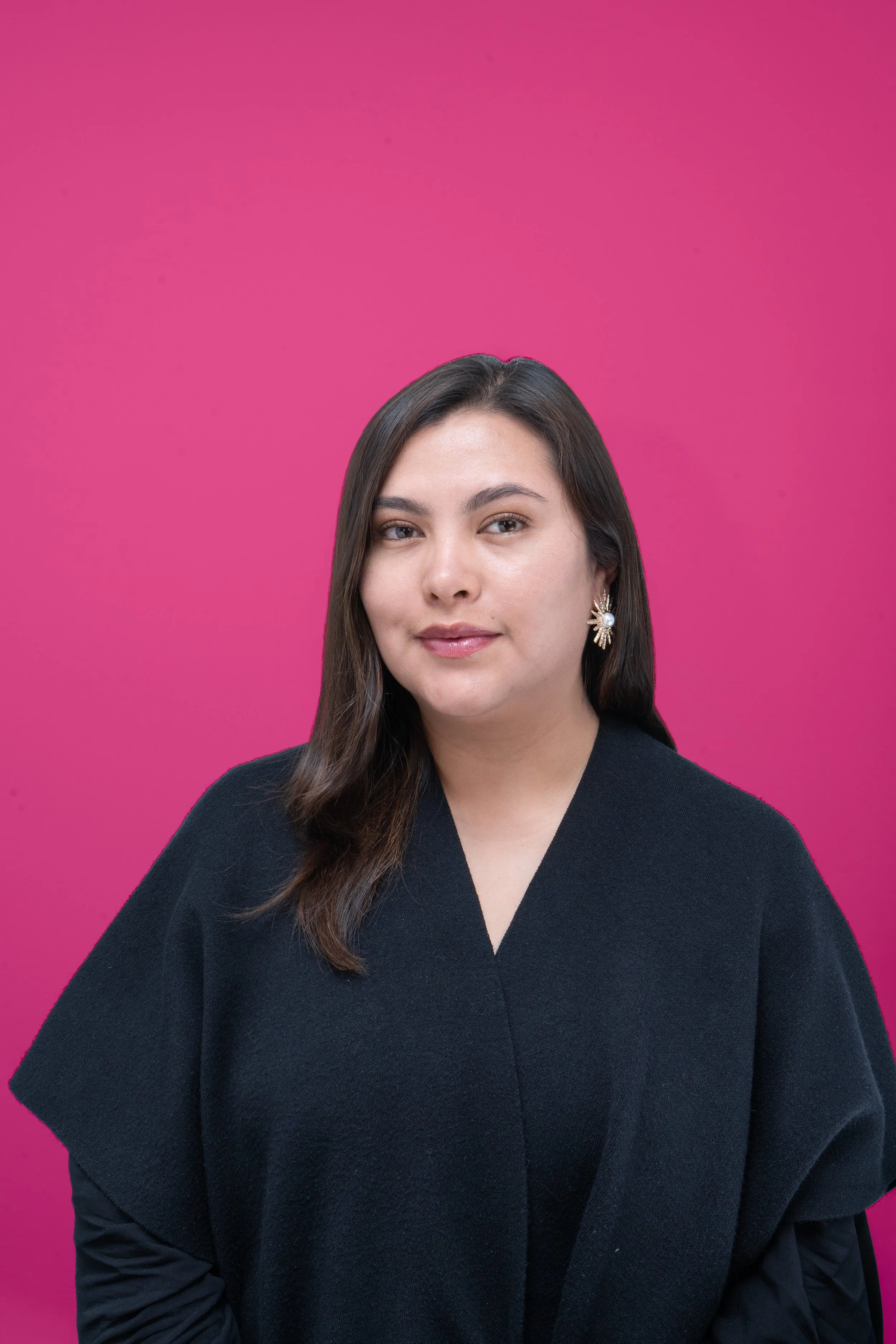 Retrato de una mujer joven con cabello castaño oscuro, cabello suelto, usando un abrigo negro y aretes decorativos, de fondo rosa.