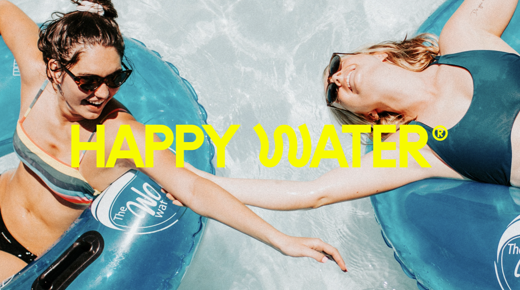 Dos mujeres disfrutando en flotadores azules en la alberca, una de ellas con gafas de sol y sonrisa, la otra con gafas de sol y cabello rubio, tomando de la mano y descansando sobre el agua.