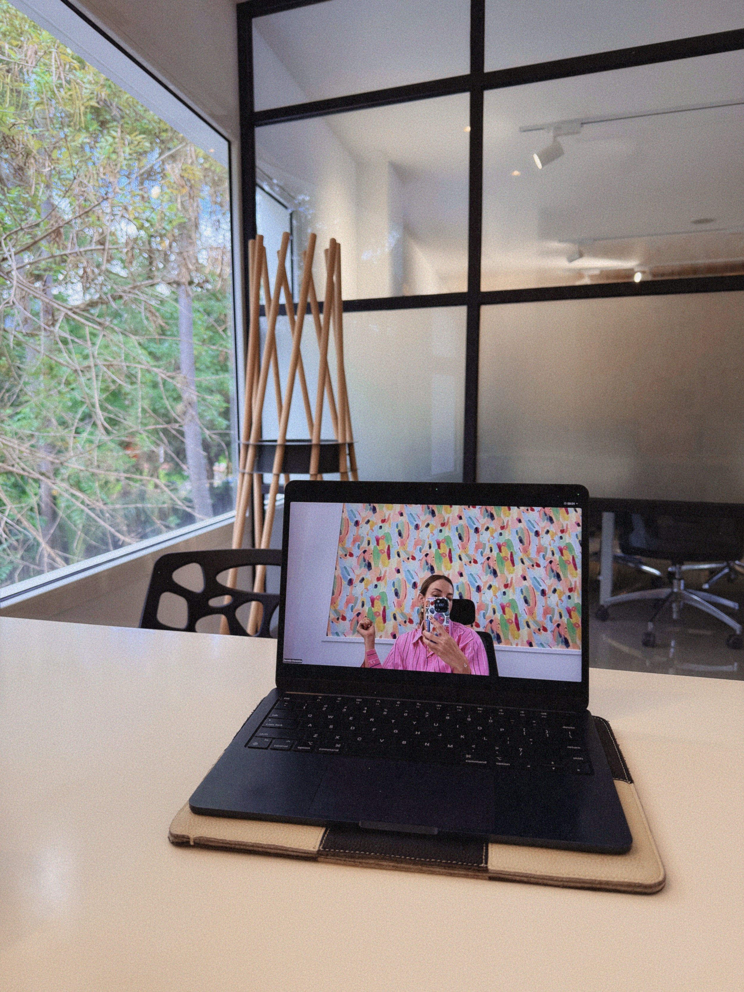 Una laptop sobre una mesa con una mujer tomándose una selfie en la pantalla, fondo de pared colorido con arte abstracto, en un ambiente de oficina moderna con ventanas grandes y una vista de árboles afuera.