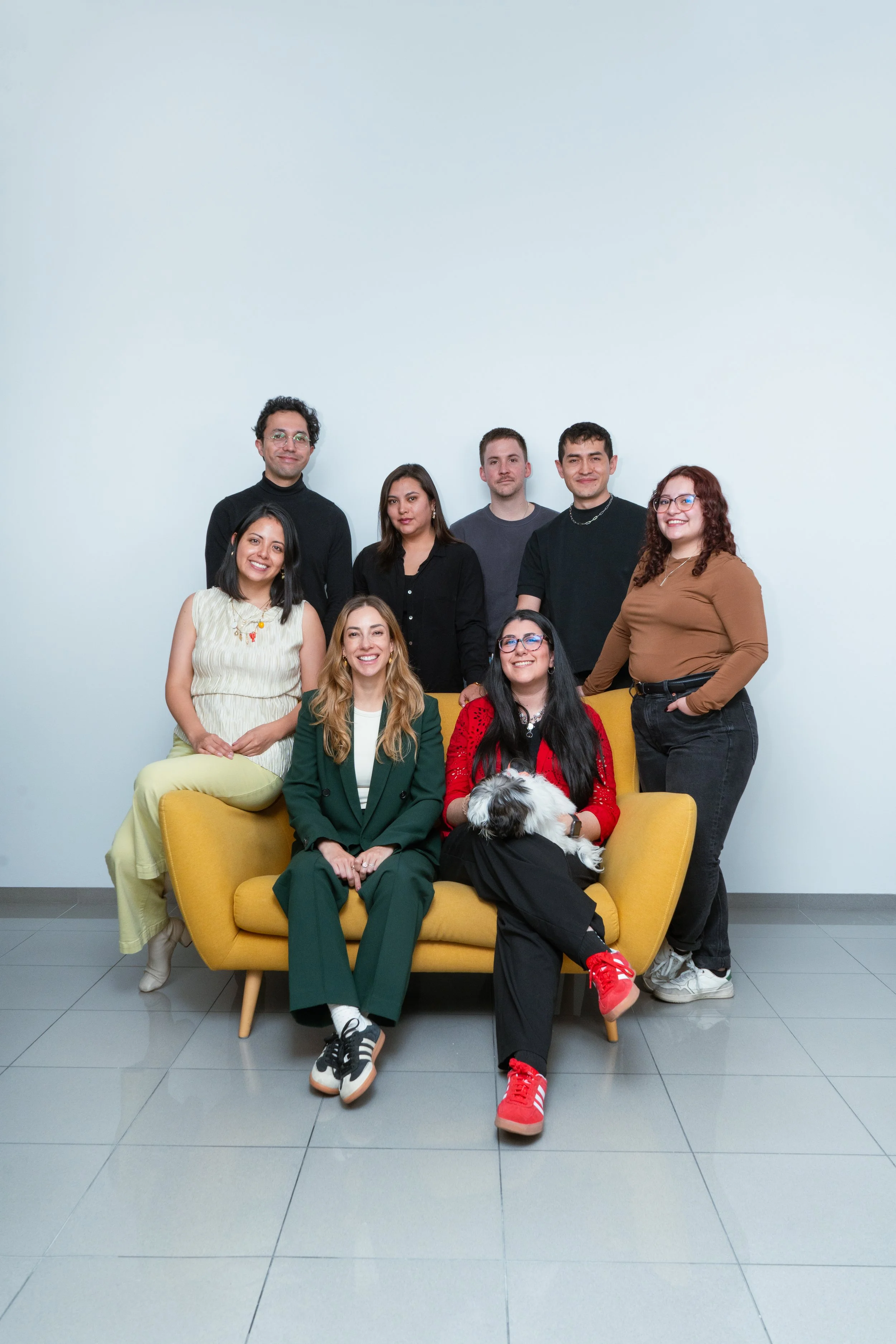 Grupo de ocho personas y un perro en un sillón amarillo, posando para una foto en una pared blanca.