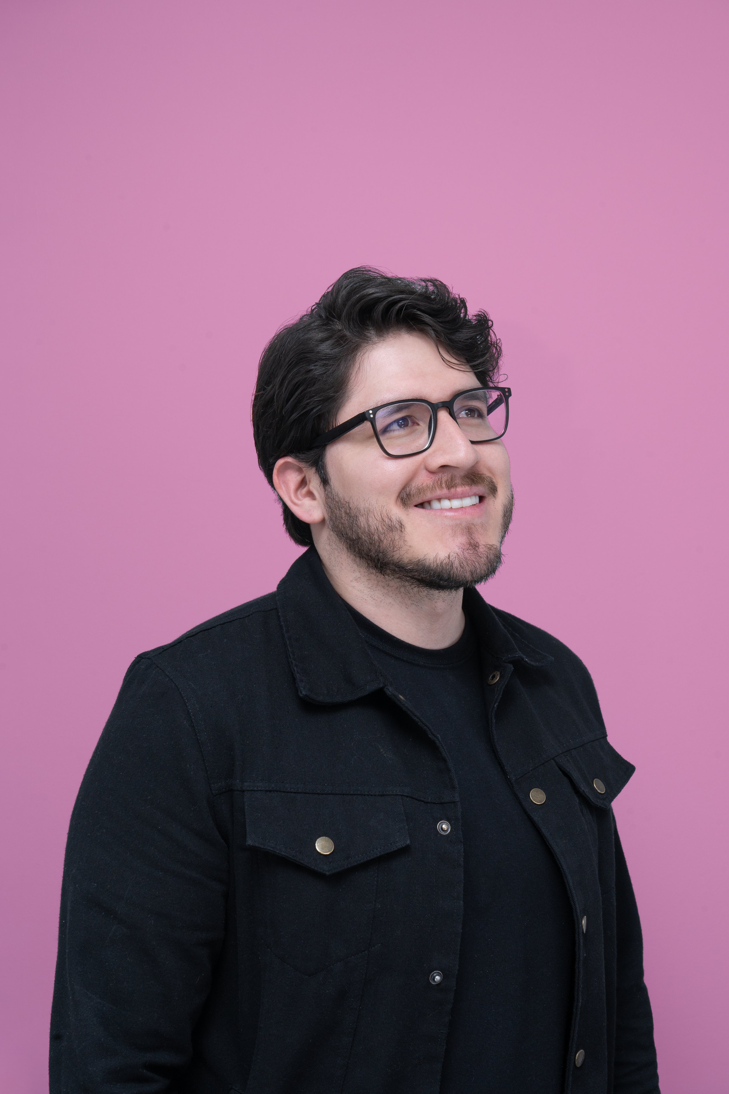 Hombre con gafas de marco negro, barba y melena oscura, sonriendo, con fondo rosa.