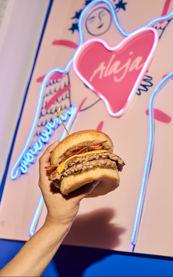 Una mano sostiene una hamburguesa con carne, tocino, queso y pan en un fondo de neón con una figura de Jesús y una palabra que dice 'Alija'.