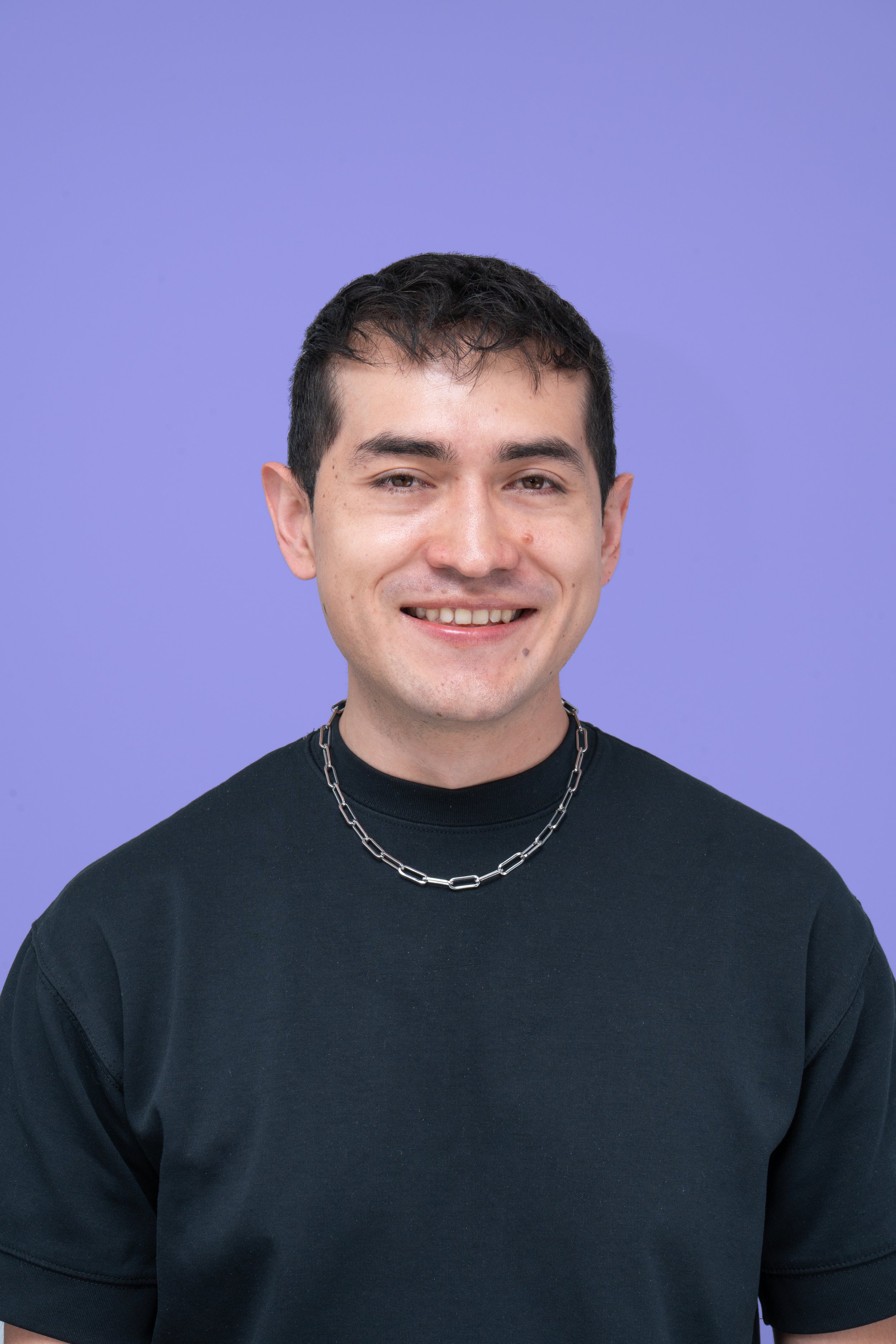Retrato de un hombre joven con camiseta negra y cadena de acero, sonriendo, fondo lila.