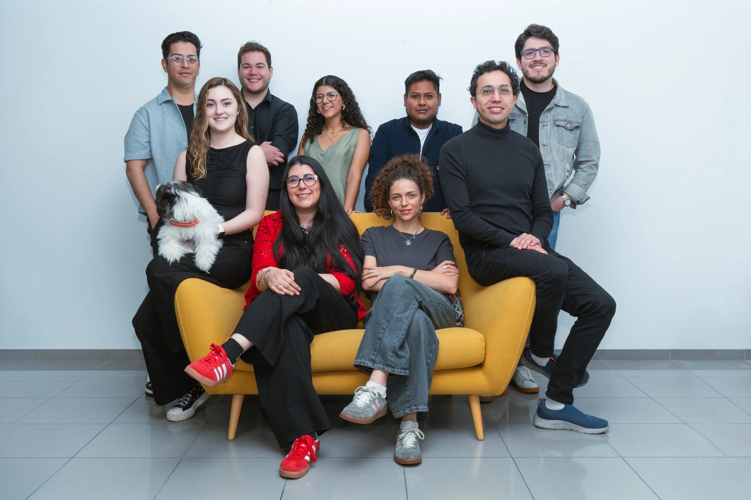 Grupo de personas posando en una sala con un sillón amarillo, incluye a una mujer con un perro blanco y negro, todos sonrientes.
