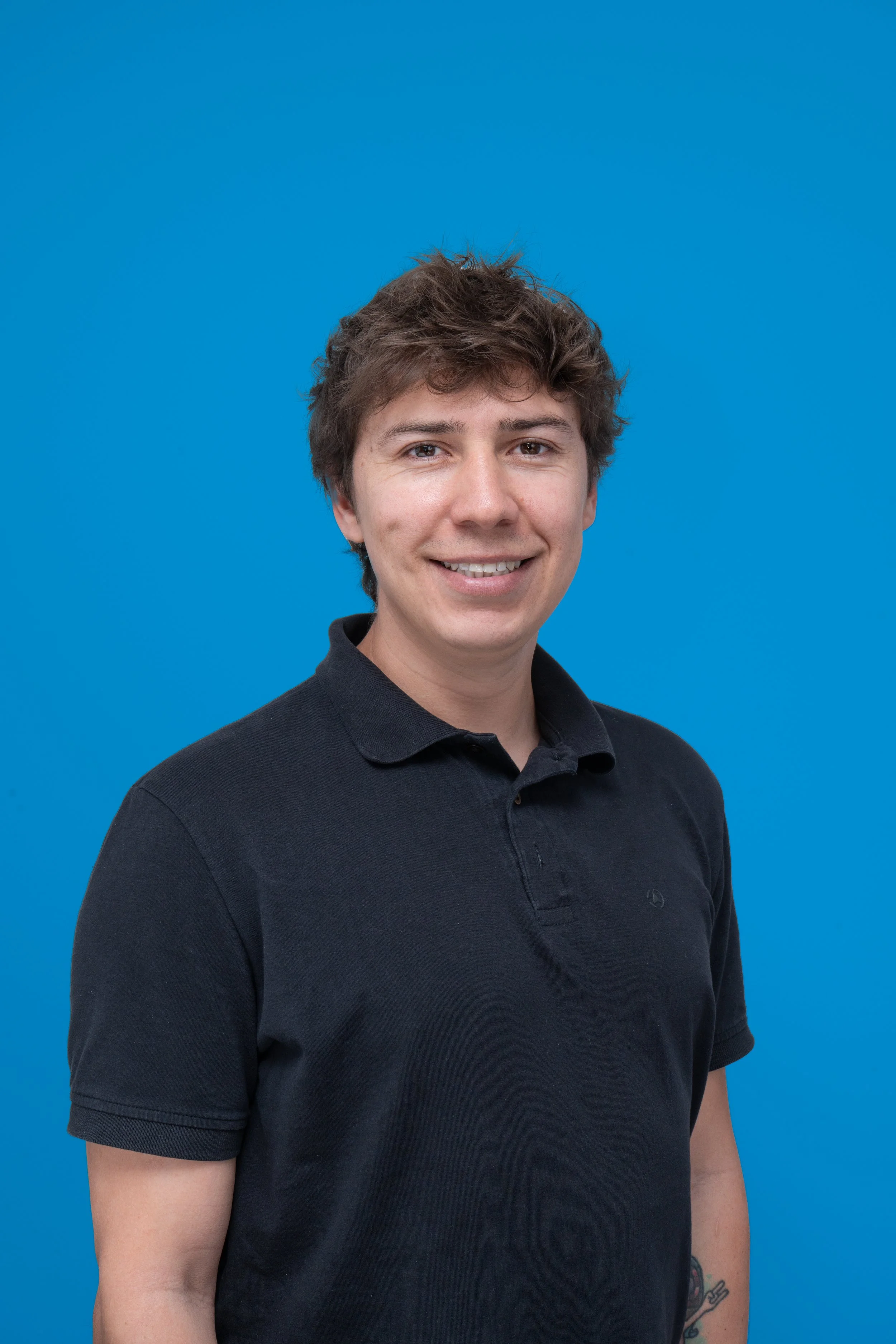 Retrato de un joven con camiseta negra, pelo castaño y peinado despeinado, sonriendo y posando frente a un fondo azul.