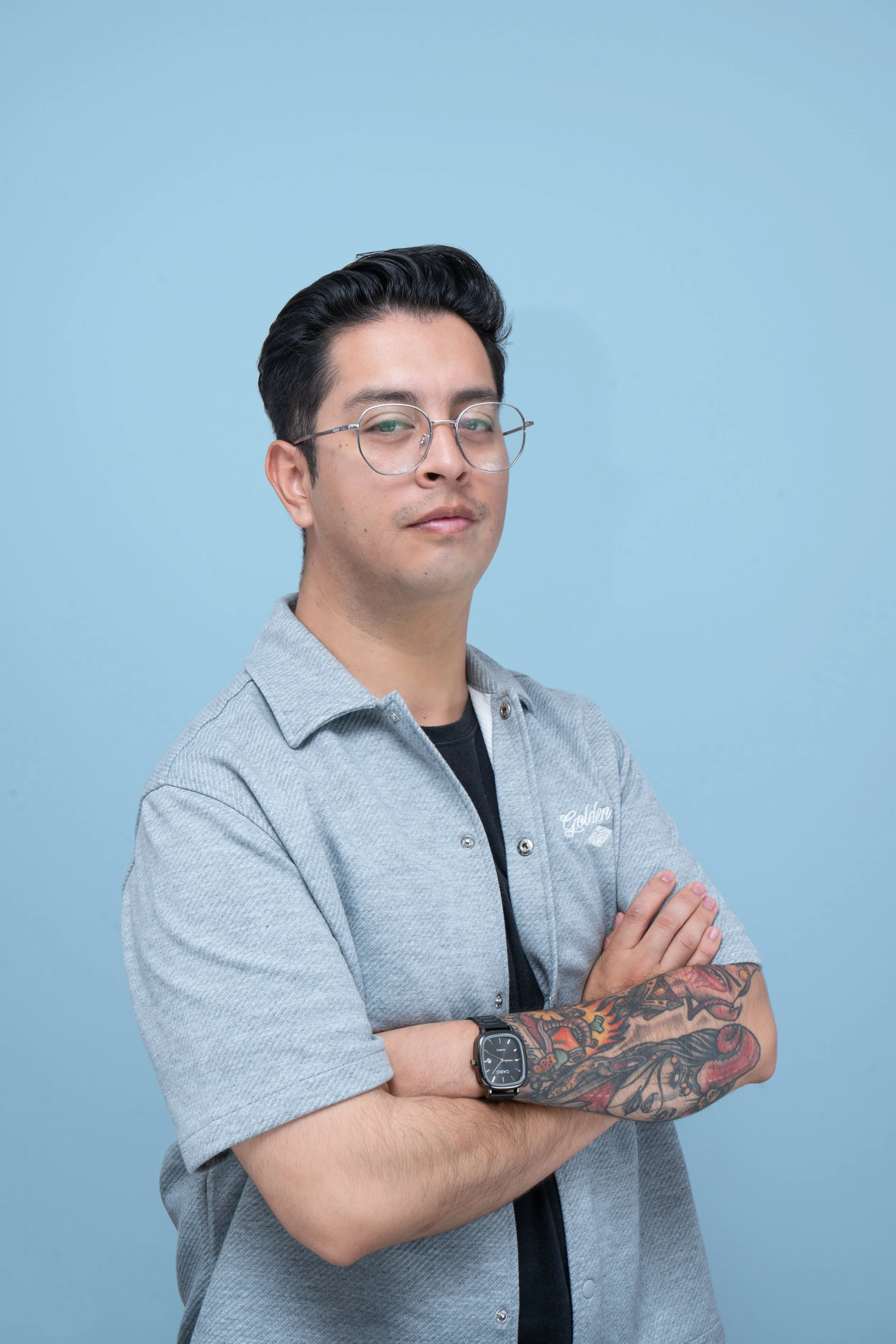 Hombre con gafas, peinado oscuro, camiseta gris con logo en el pecho, brazo tatuado con dragón y reloj en la muñeca, cruzando los brazos, fondo azul.