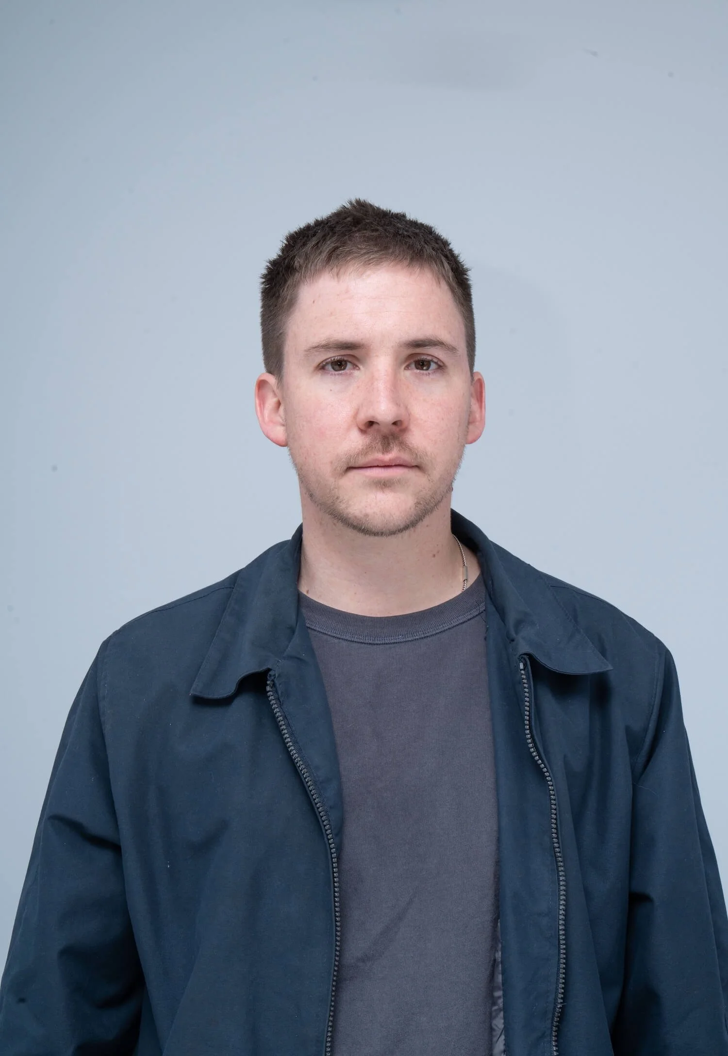 Joven hombre con camiseta gris y chaqueta azul, pelirrojo y con barba, mira hacia la cámara con fondo gris.