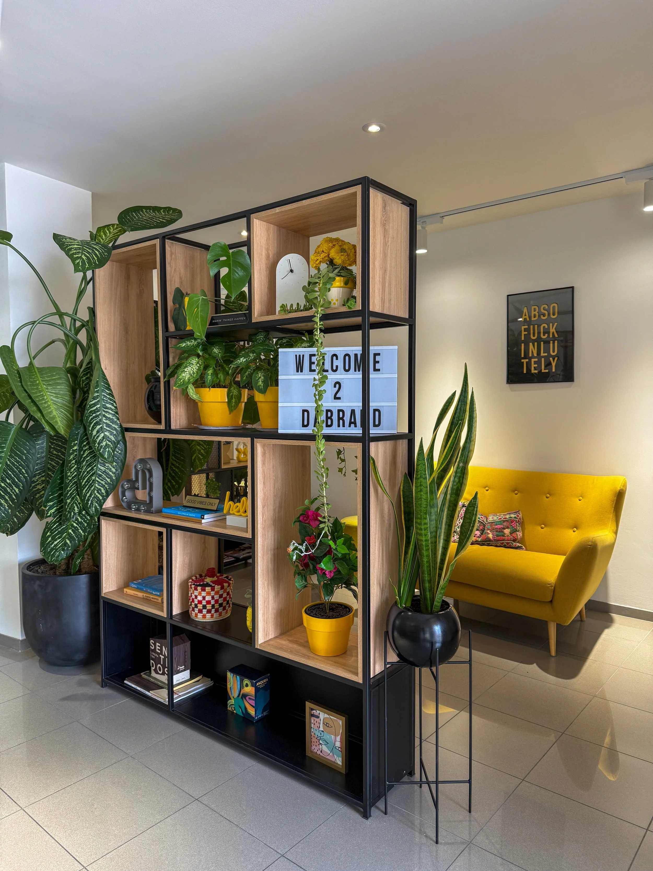 Estantería con plantas verdes y decoraciones en un espacio interior, con un sillón amarillo en el fondo y un cartel que dice 'ABSOLUTELY INFLUENTLY' en la pared.