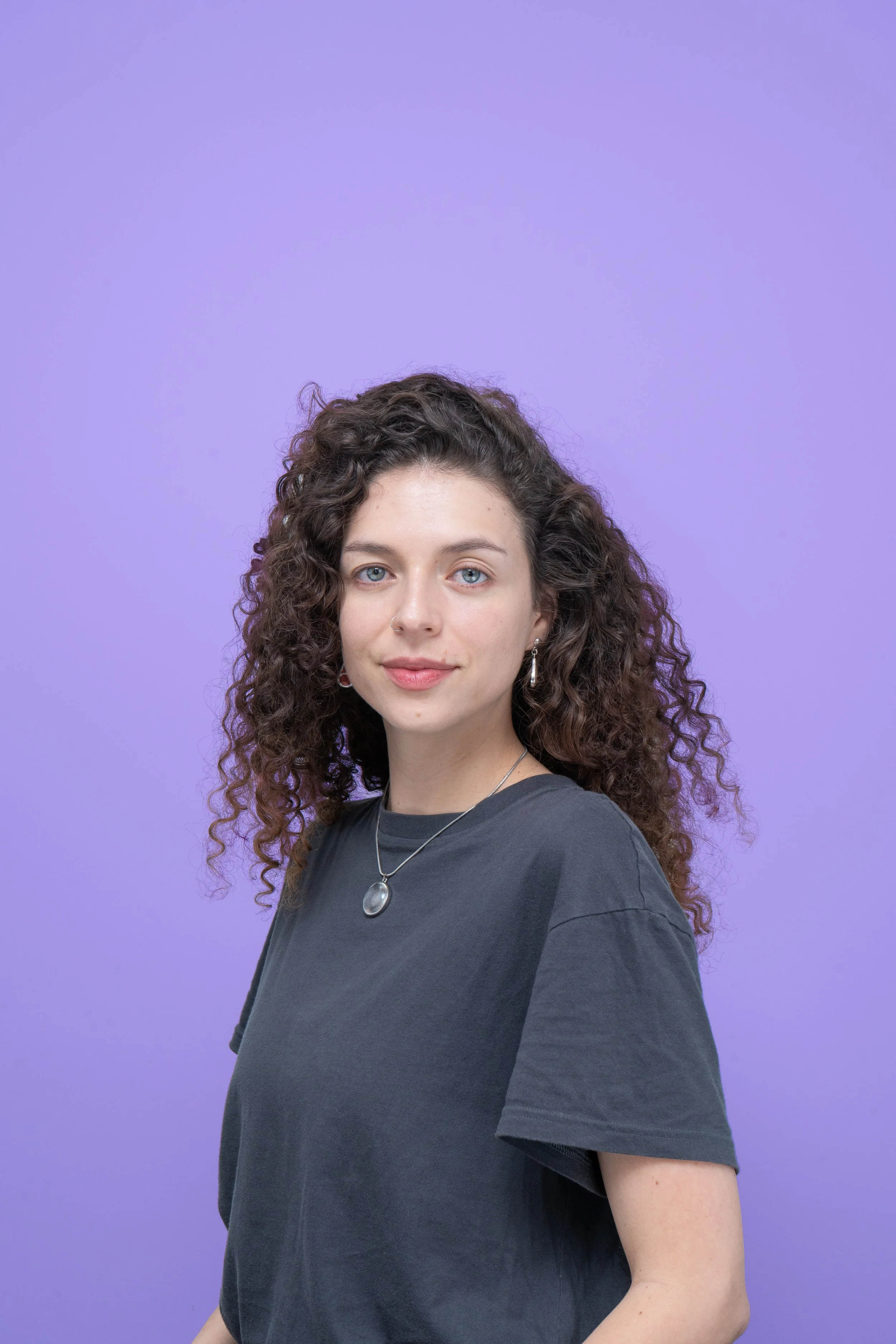 Joven mujer con cabello rizado, vestida con camiseta gris oscuro, usando collar y pendientes, frente a un fondo morado.