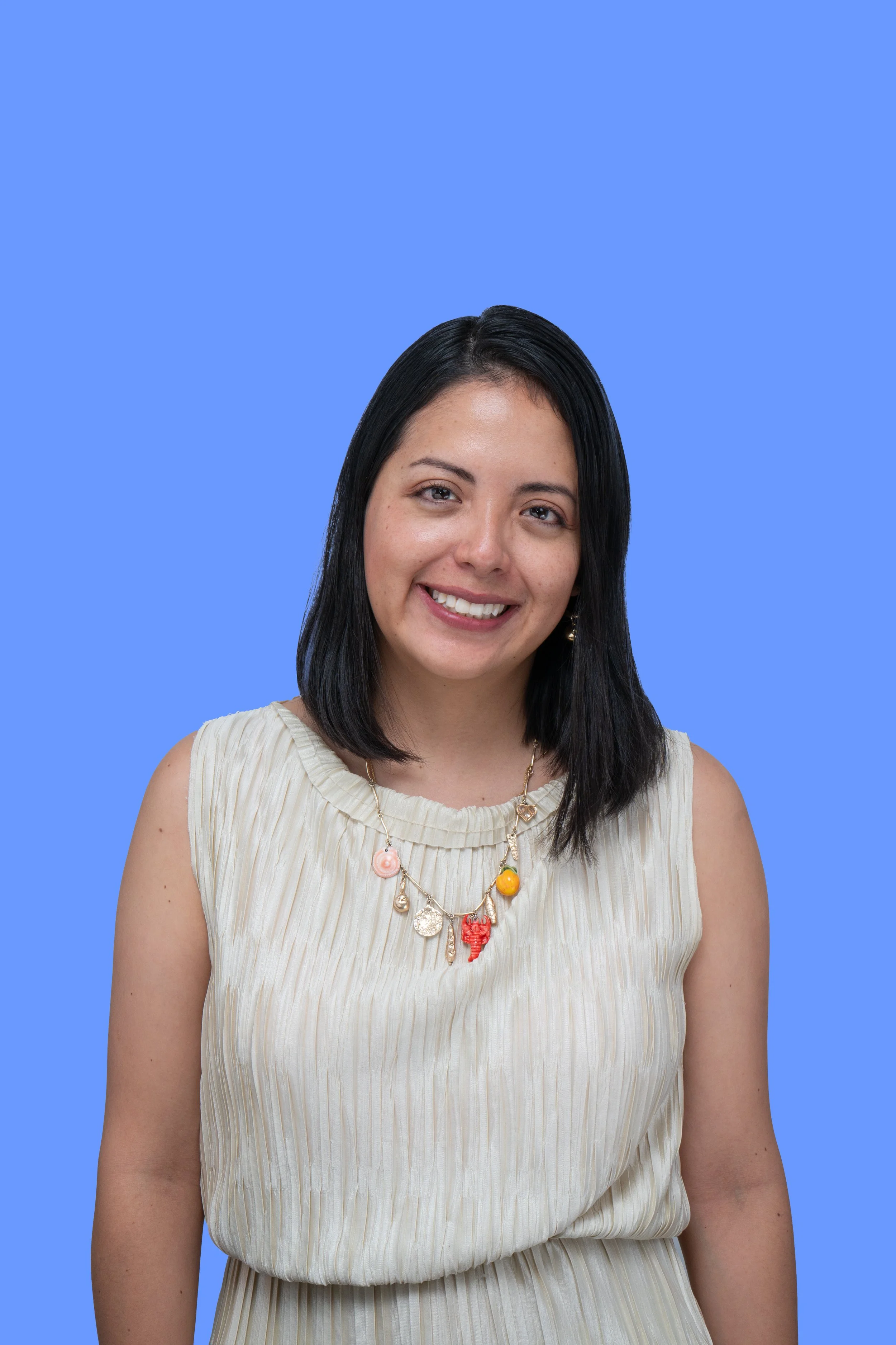 Retrato de una mujer con cabello negro, camiseta beige y collar colorido, sonriendo frente a un fondo azul