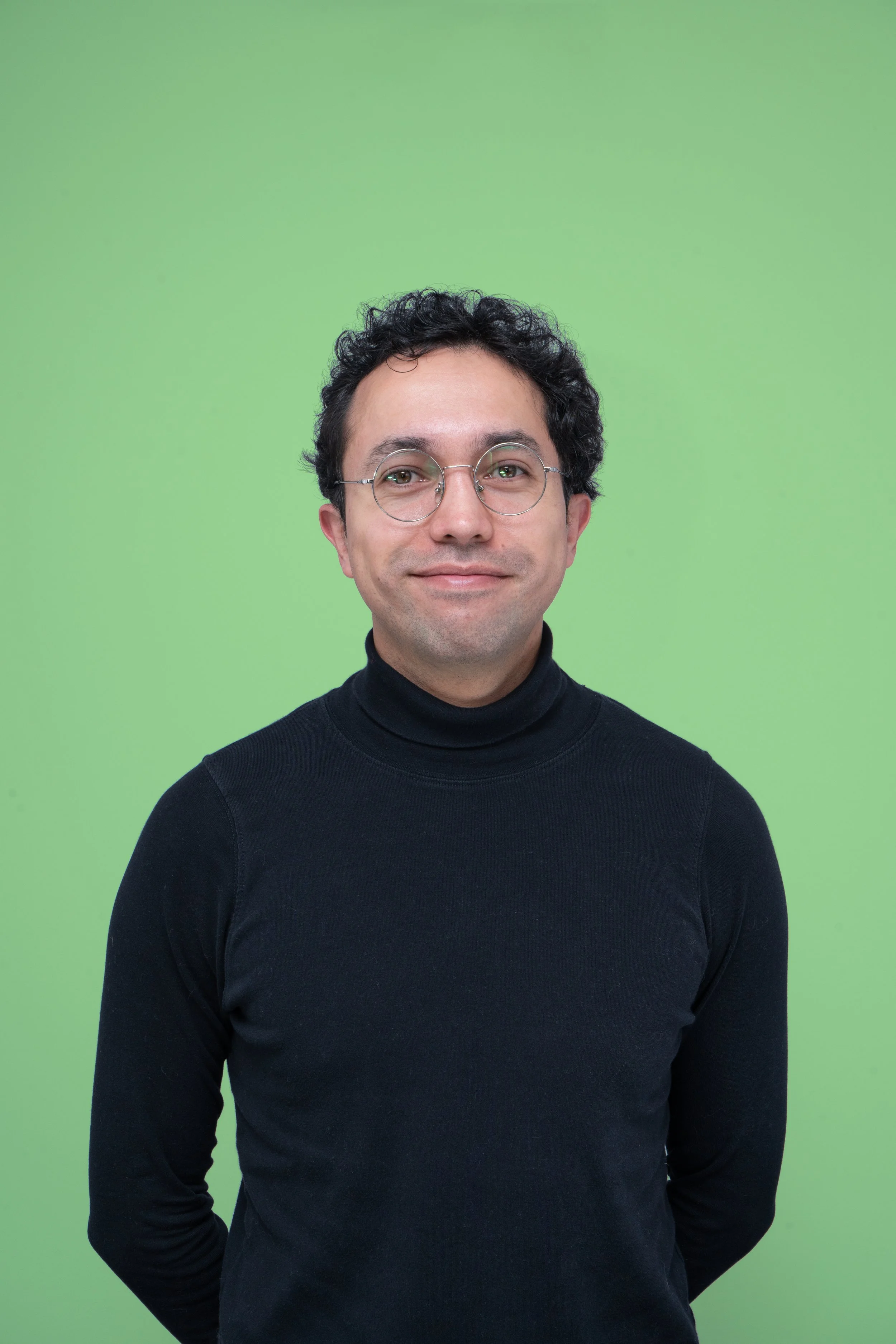 Hombre con gafas, cabello rizado y oscuro, usando suéter negro, sonriendo ligeramente, fondo verde liso.