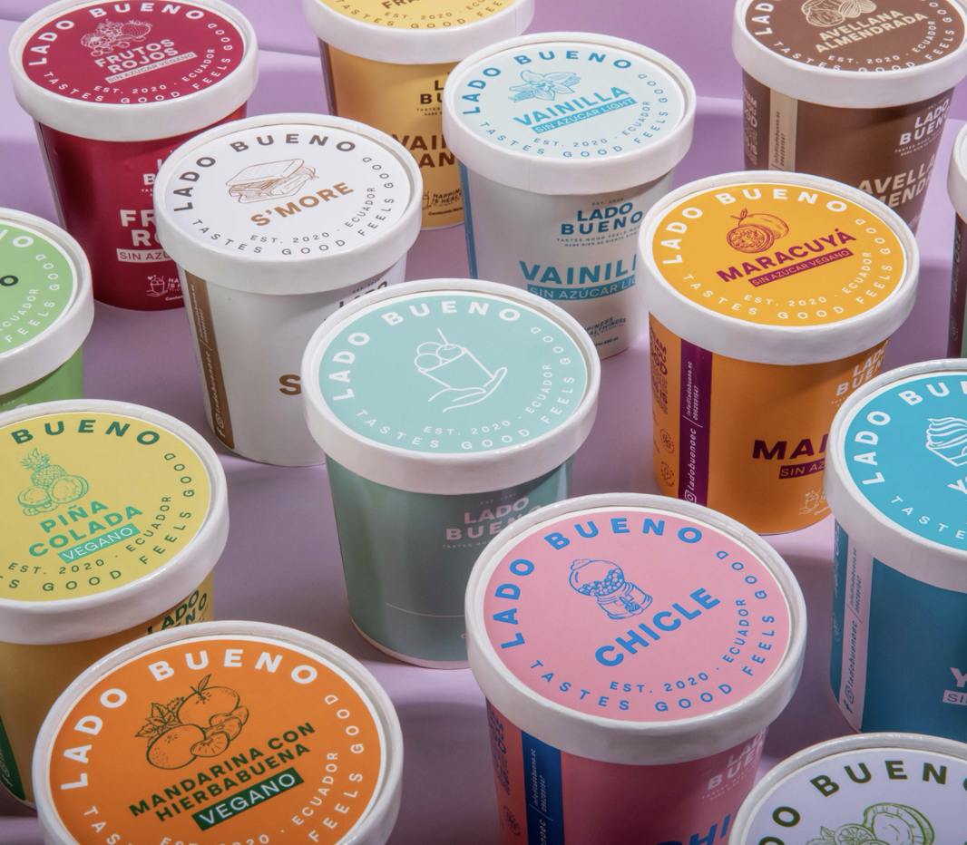 Conjunto de varios envases de helado de sabores diferentes, con colores vivos y etiquetas que indican los sabores como fruta roja, vainilla, maracuya y chicle, sobre un fondo rosado.