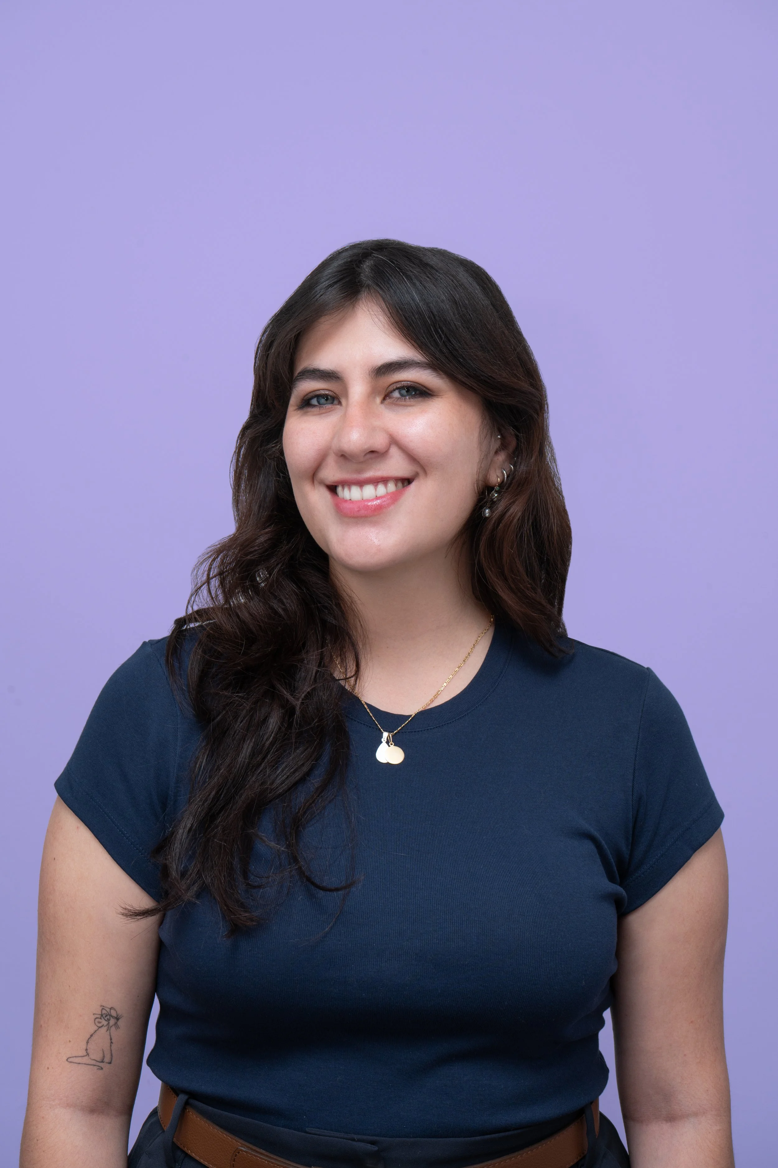 Retrato de una mujer sonriendo con cabello castaño oscuro, llevo camiseta azul y collar dorado, fondo morado claro.