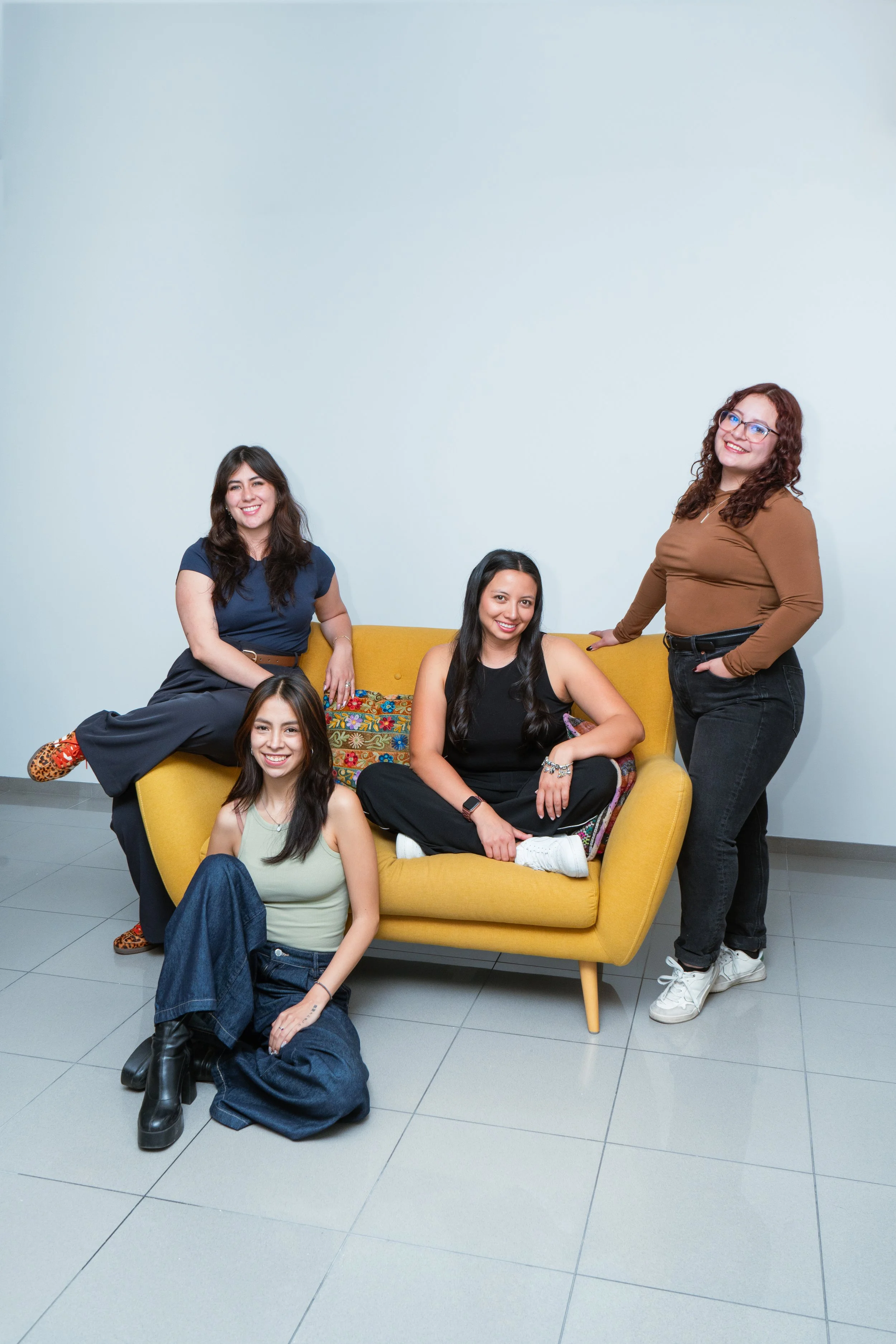 Cinco mujeres sonrientes en un ambiente interior, con una de ellas sentada en el suelo y las otras cuatro en un sillón amarillo.