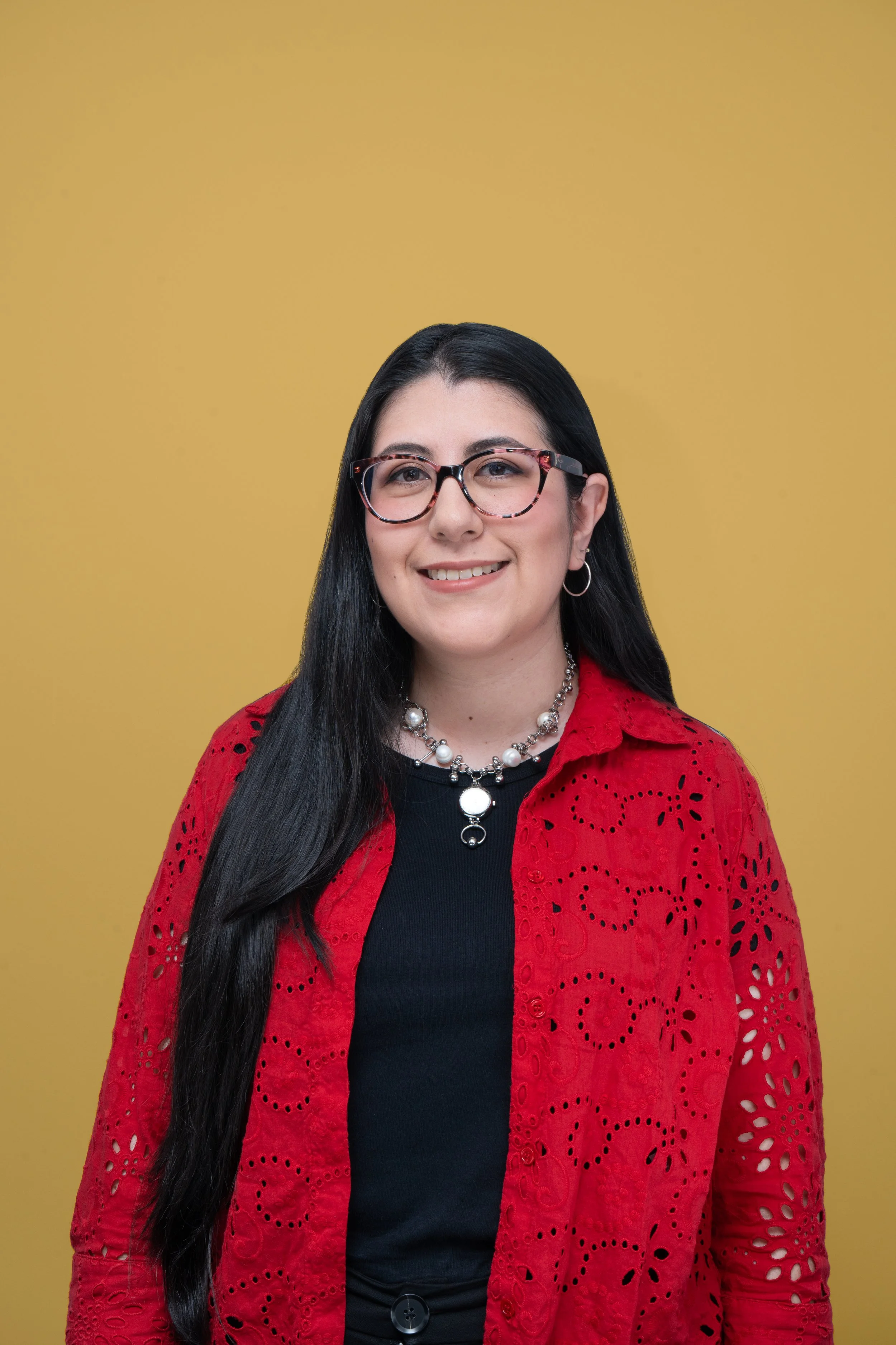 Retrato de una mujer de cabello largo y negro, luciendo gafas con marco rosa y negro, acampanada y con detalles en blanco, usando un collar de perlas y aretes pequeños en aro, vestida con una camiseta negra y una chaqueta roja con detalles perforados