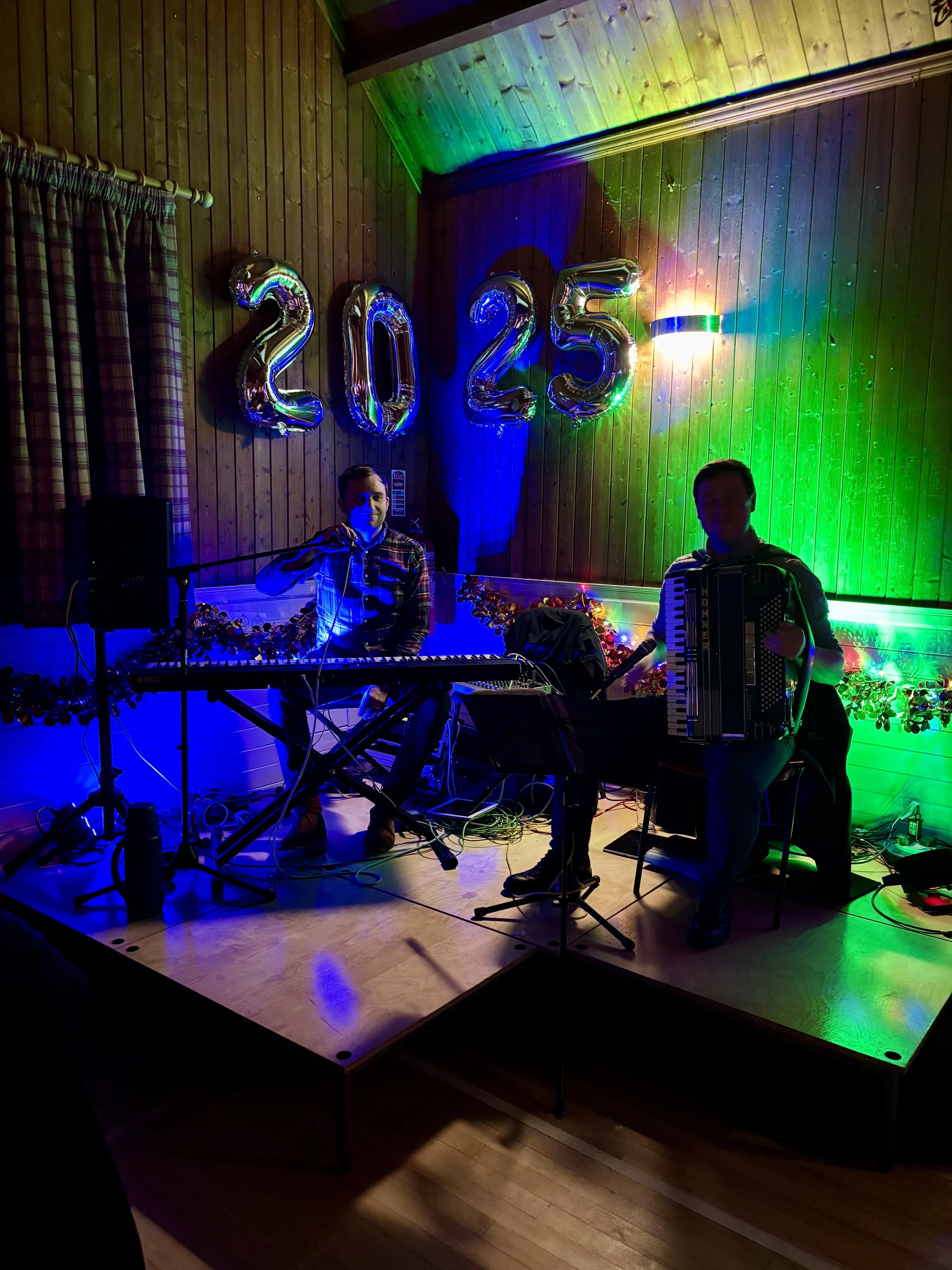 Two musicians performing on a wooden stage decorated with colorful lights and large silver balloons spelling '2025'. One plays a keyboard and the other plays an accordion. The scene has festive multicolored lighting.