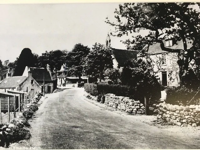 A black-and-white photo of a rural village street with houses, trees, and a stone wall, with a winding road in the foreground.