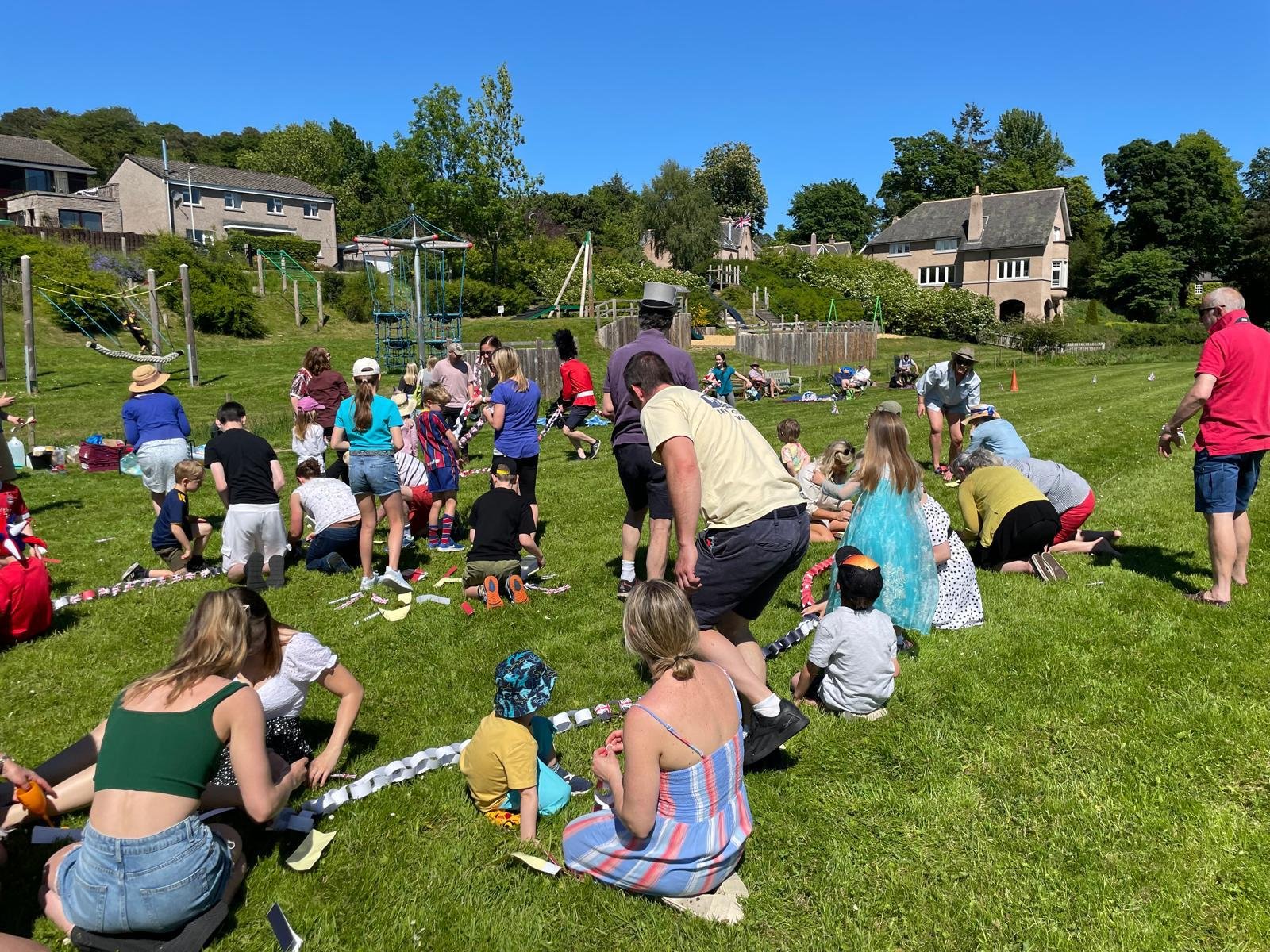 A group of children and adults gathered on the grass at Strachan Park playing field in Feughside, at a community event, with houses and trees in the background.