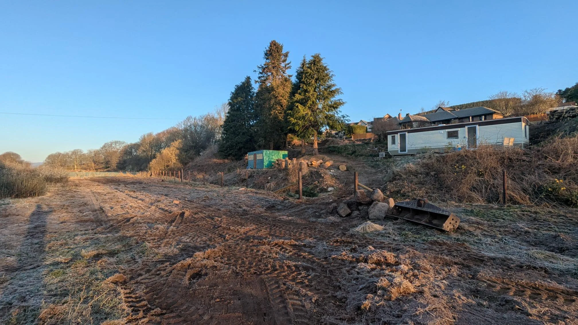 Feughside/Strachan Community Garden. A dirt road with tire tracks, a fence on the side, and a hillside with trees and mobile homes or cabins on the slope.